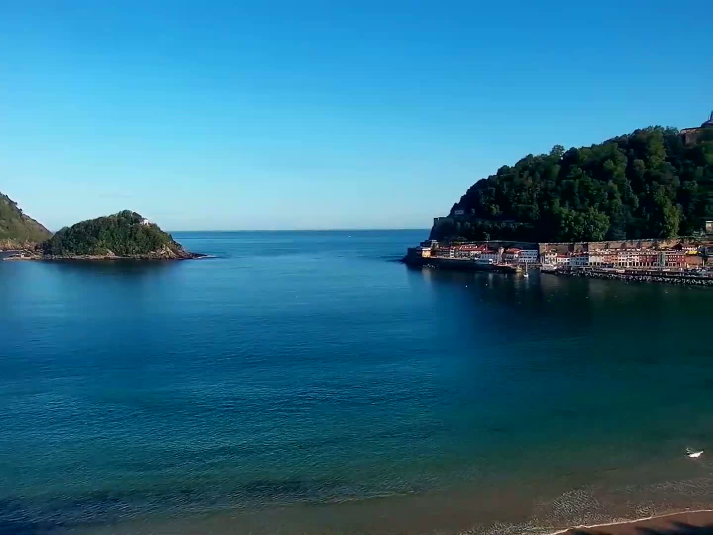 A tranquil, clear blue ocean separates two verdant hillsides, one featuring a small coastal town.