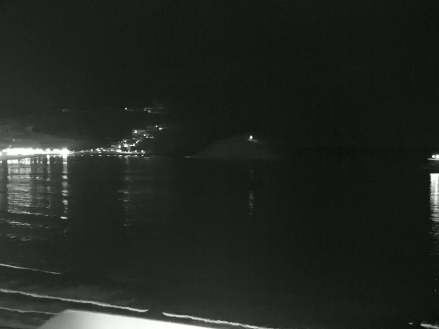 A nighttime view of a calm ocean reflecting city lights on the shore and a small island in the distance.