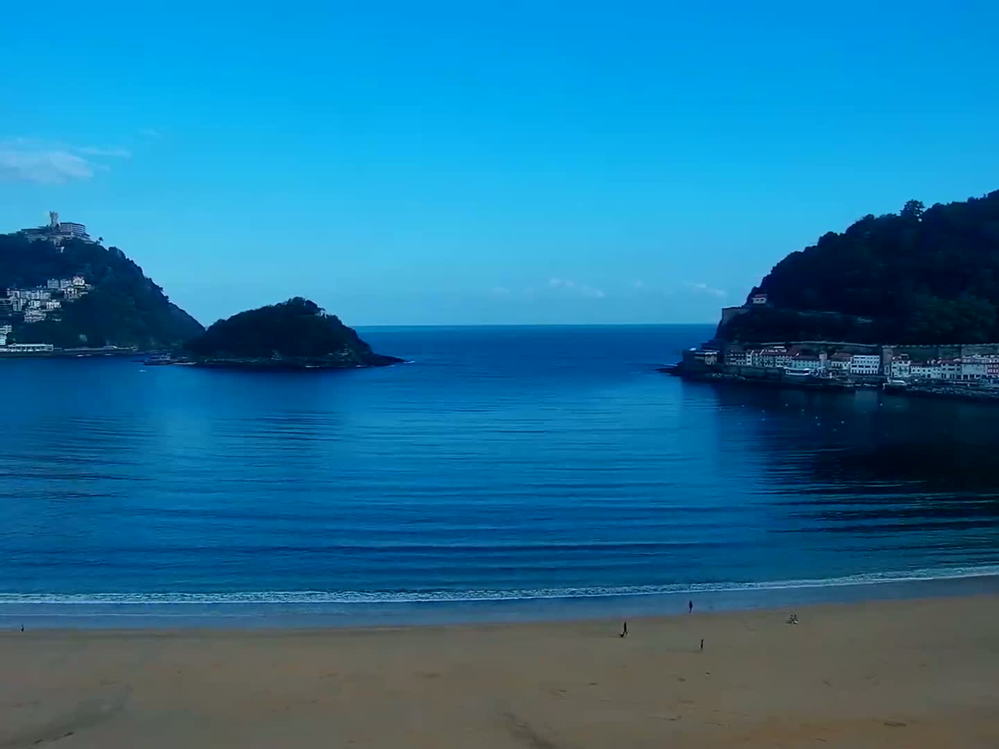 A sandy beach stretches beneath a clear blue sky, facing a calm blue sea separating two hillsides with buildings.