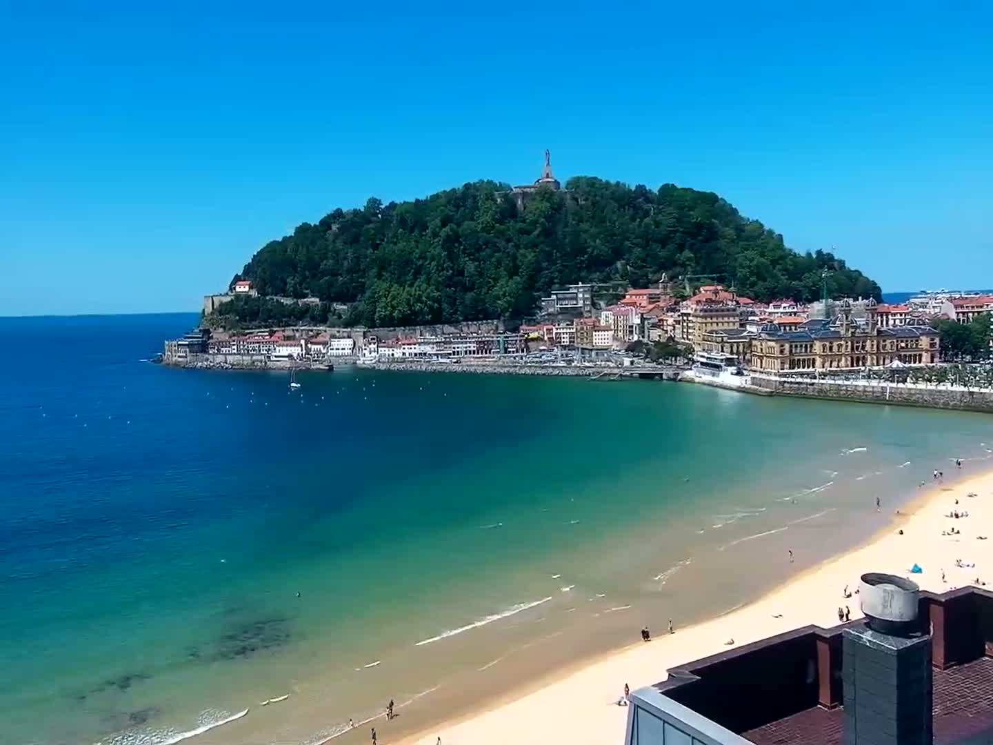 A sandy beach and calm, blue-green ocean meet a town nestled at the base of a lush, green hill under a clear, bright blue sky.