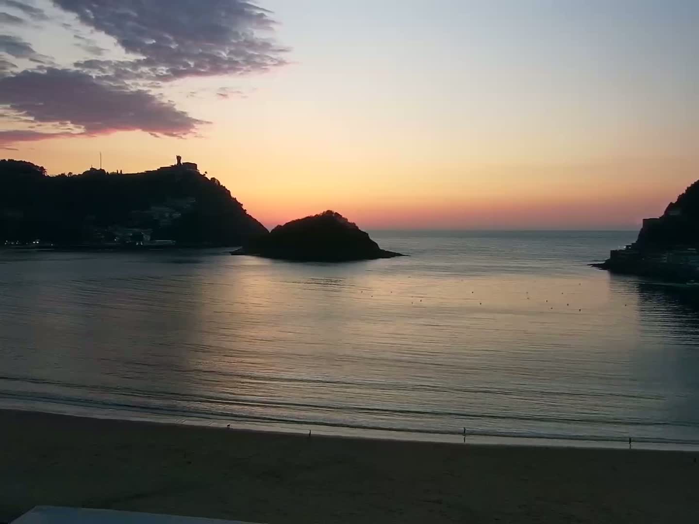 A calm bay at sunset, with two silhouetted hills on either side of a sandy beach.