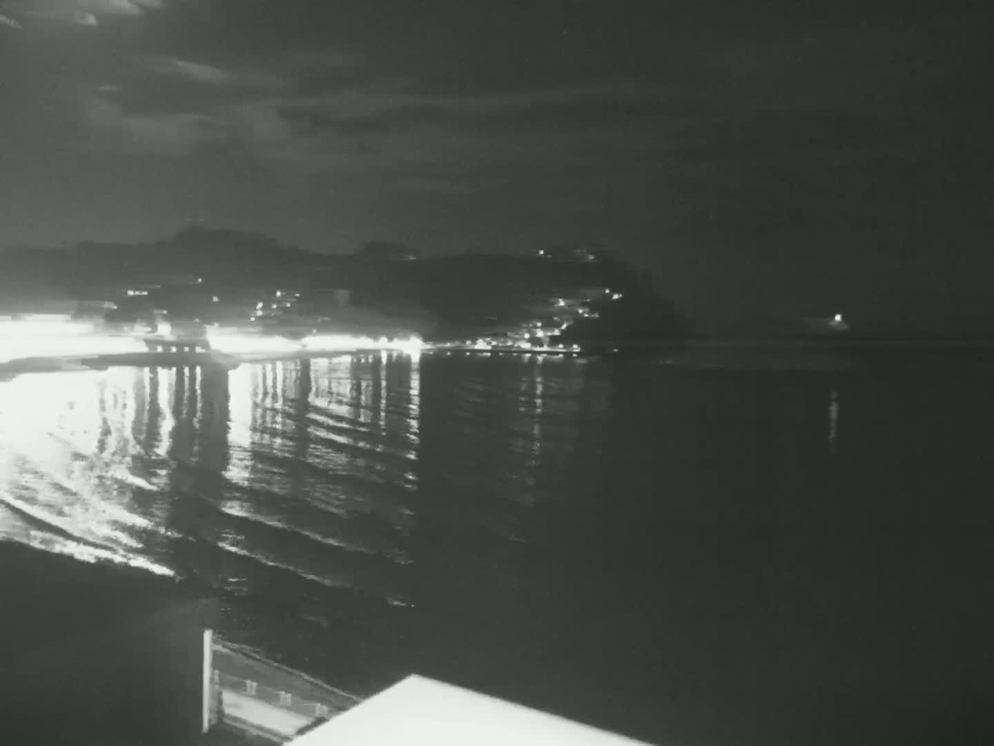 A nighttime view of a calm body of water reflecting the lights of a town on a hillside under a partly cloudy sky.
