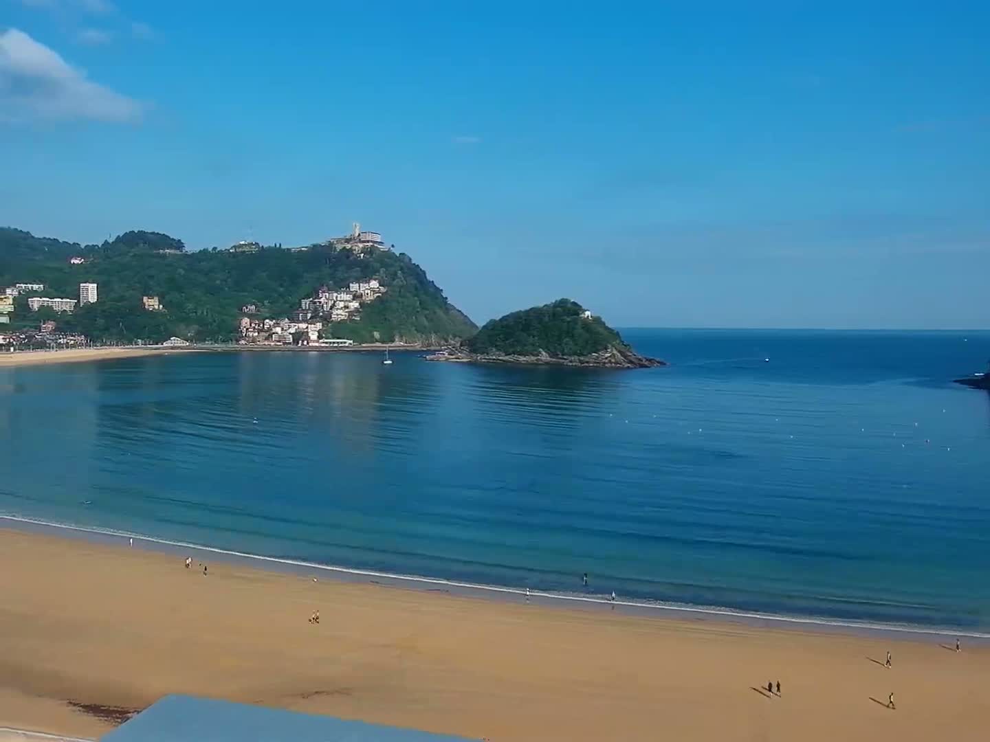 A sandy beach with a few people on it stretches along a calm, blue bay, backed by a green hill dotted with buildings under a clear, sunny sky.