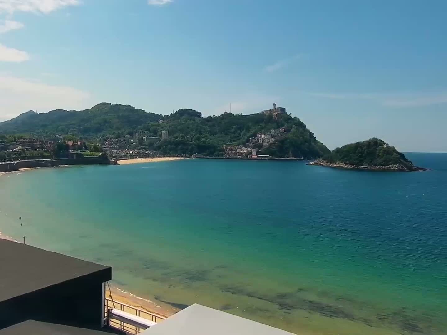 A sunny day reveals a coastal scene with a sandy beach, clear teal water, and a verdant hill topped with buildings in the distance.