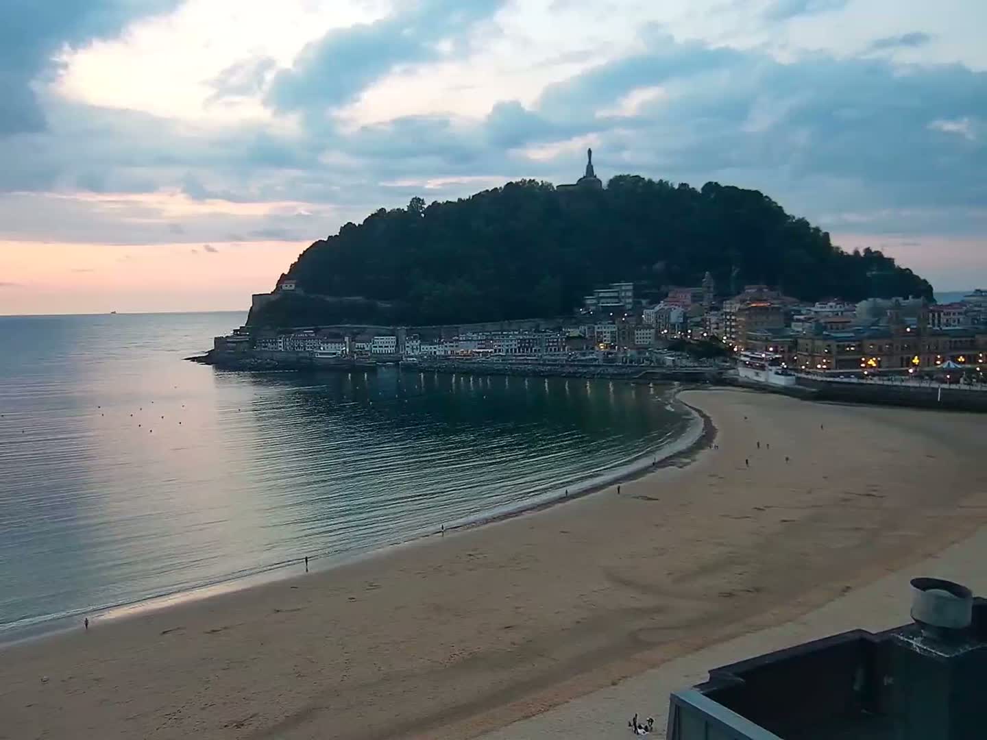 A sandy beach curves around a calm bay at sunset, next to a town nestled at the base of a verdant hill.