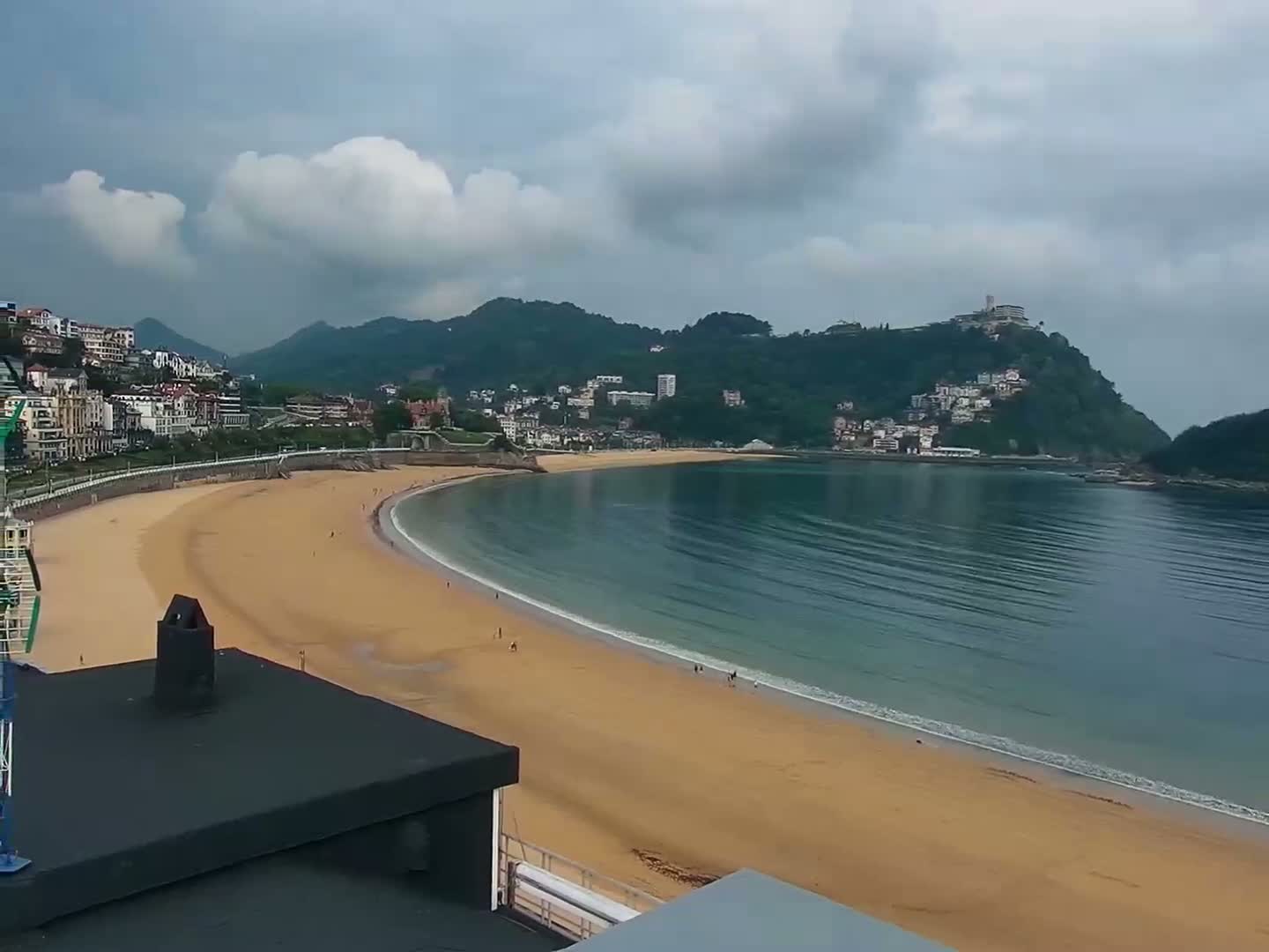 A sandy beach curves around a calm bay, backed by a town nestled against green hills under a cloudy sky.