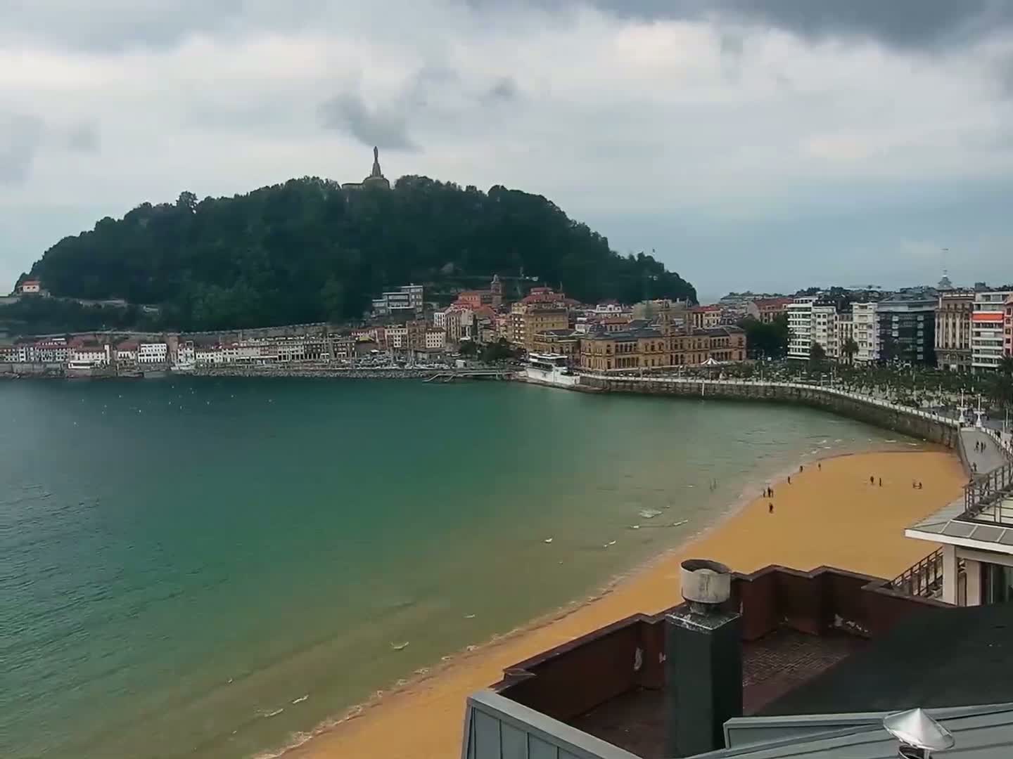 A sandy beach curves around a bay of teal water, facing a town nestled at the base of a green hill topped with a monument, all under a cloudy sky.