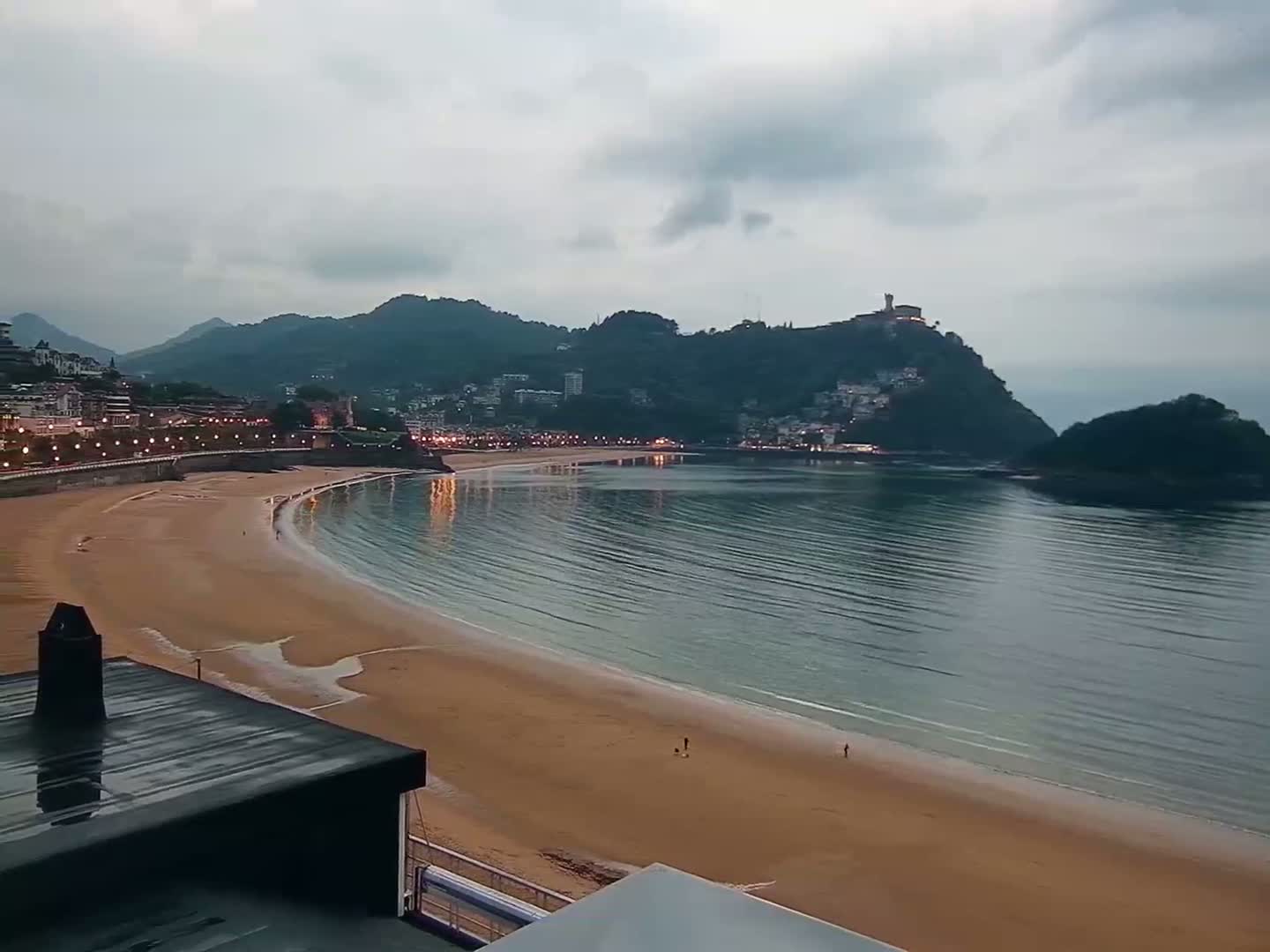 A sandy beach curves around a calm bay under a cloudy sky, with a town and hilltop buildings visible in the distance.