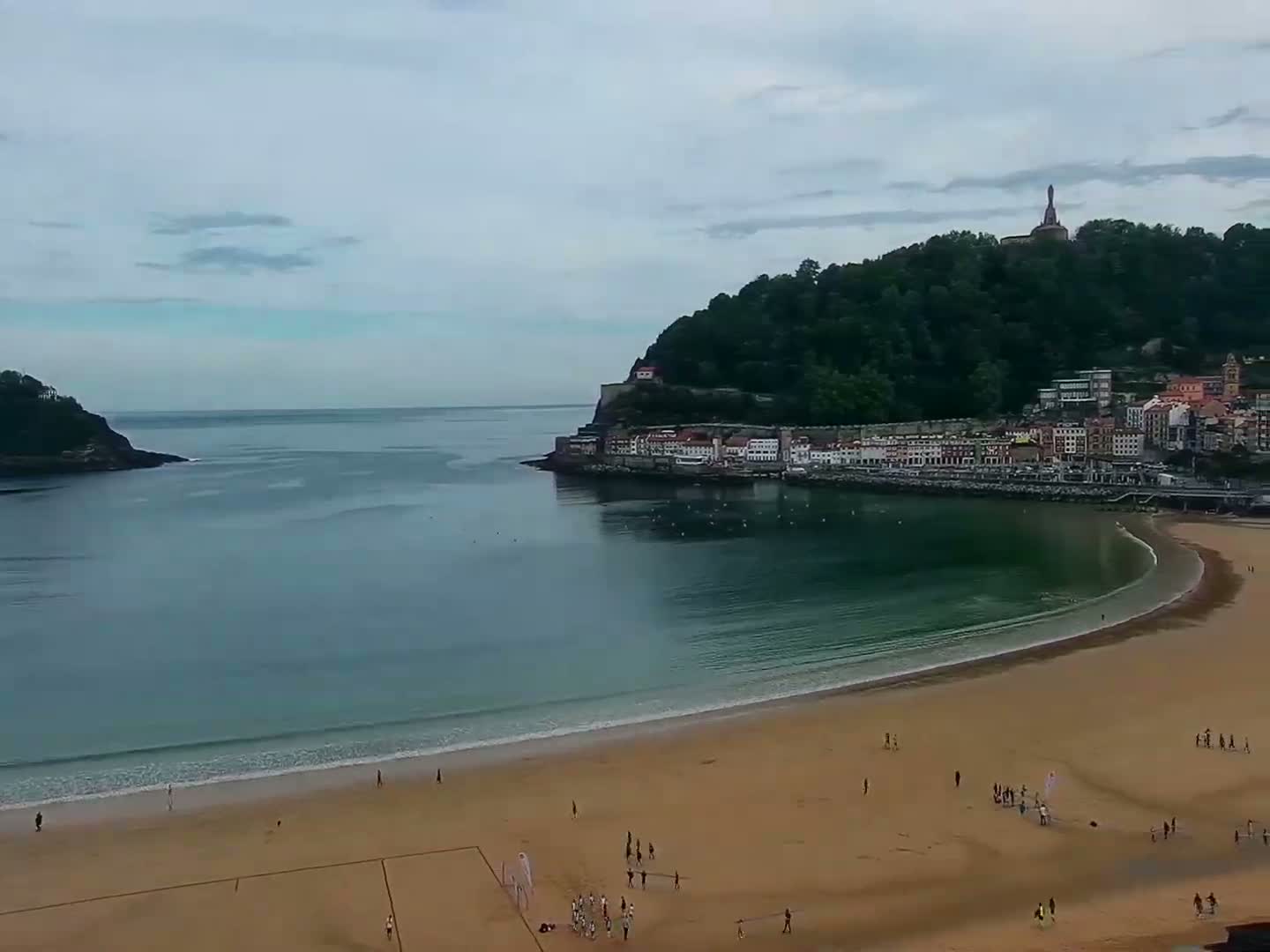A sandy beach curves around a calm bay, bordered by a town nestled against a verdant hill under a cloudy sky.