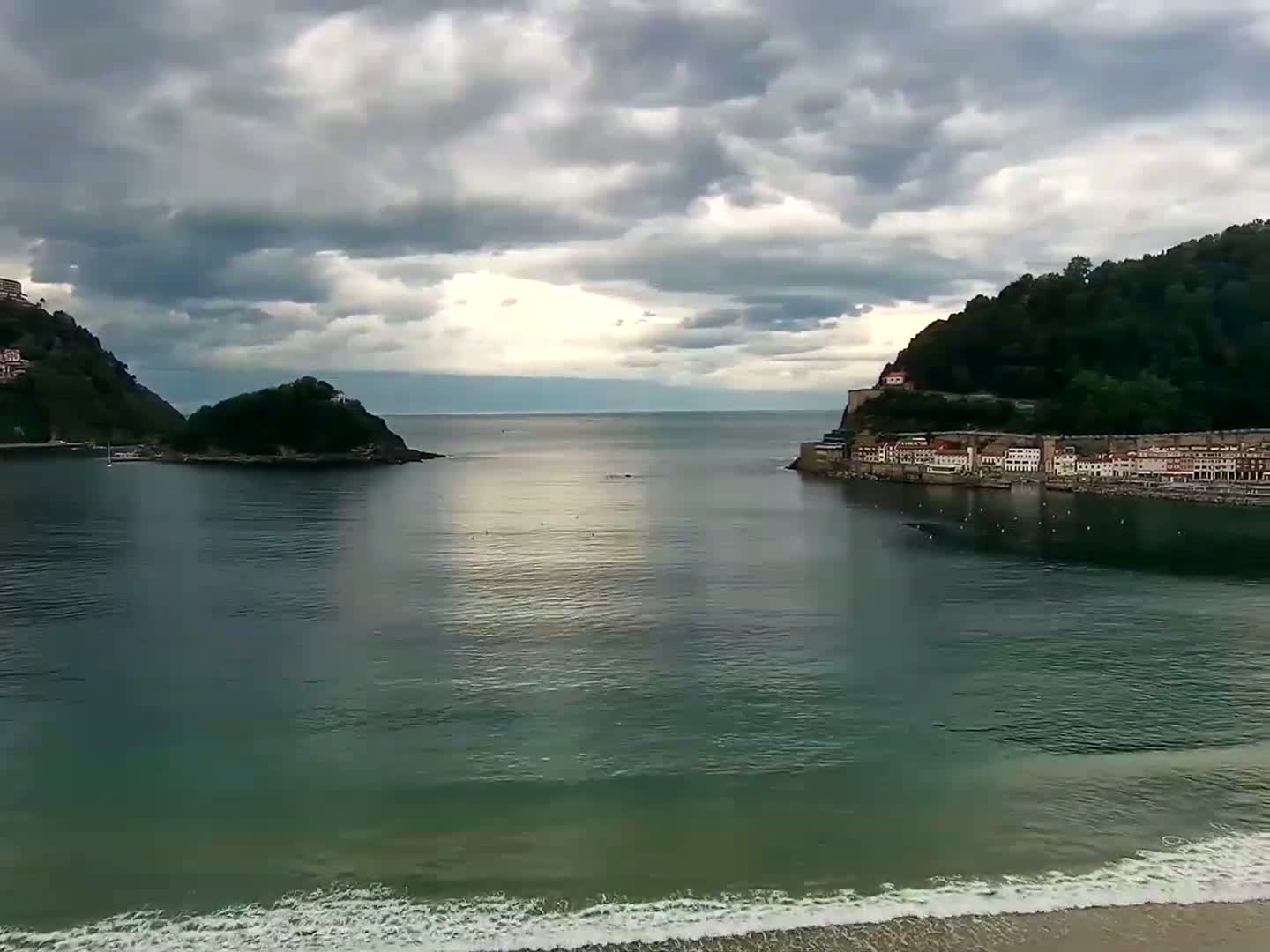 A calm bay with a small island and a hillside town under a cloudy sky, small waves lapping at a sandy beach.