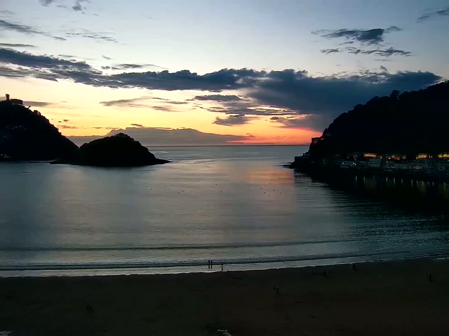 A tranquil beach scene at sunset shows two dark, rocky hills on either side of a calm bay, with a few people walking along the sandy shore.