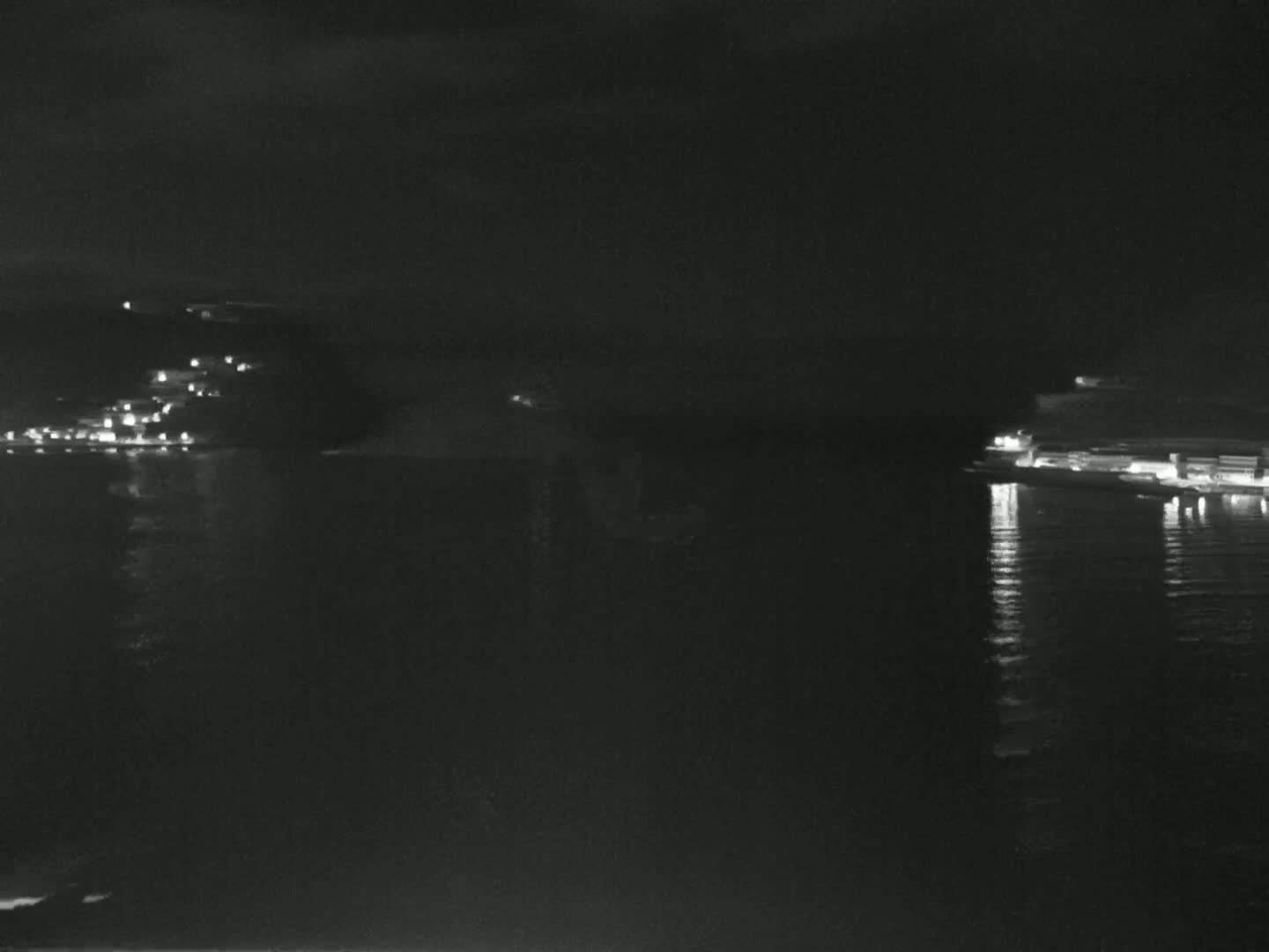 A nighttime, long shot shows a dark body of water reflecting lights from buildings on either shore under a dark sky.