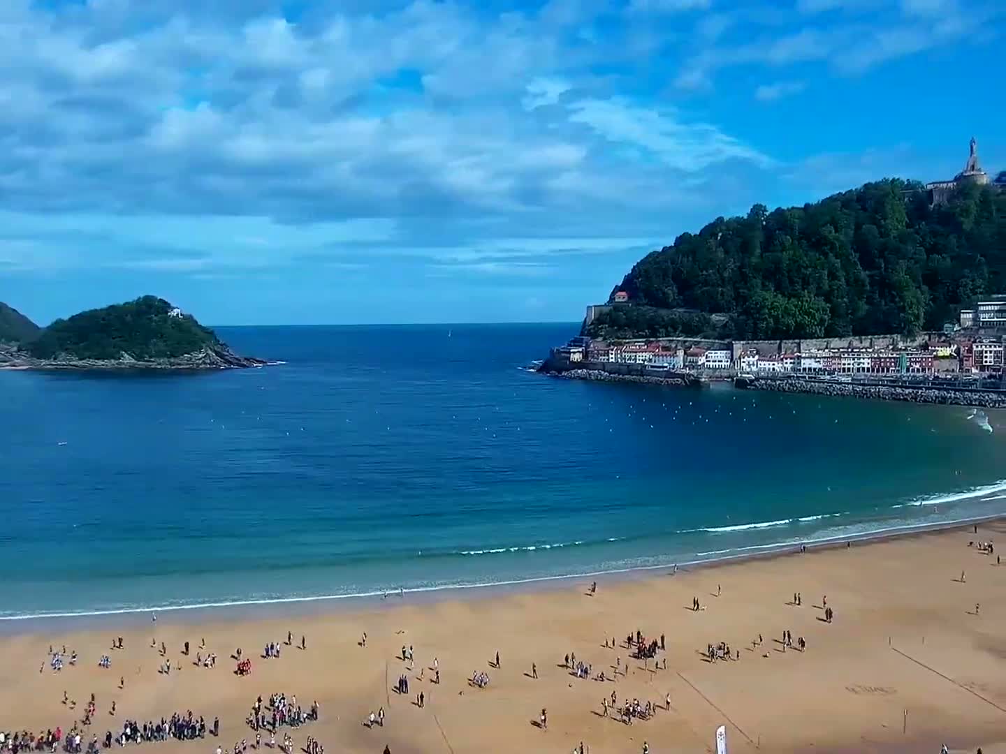 A sandy beach with many people on it is nestled in a cove between two verdant hills under a partly cloudy sky, the blue water extending to a distant island.