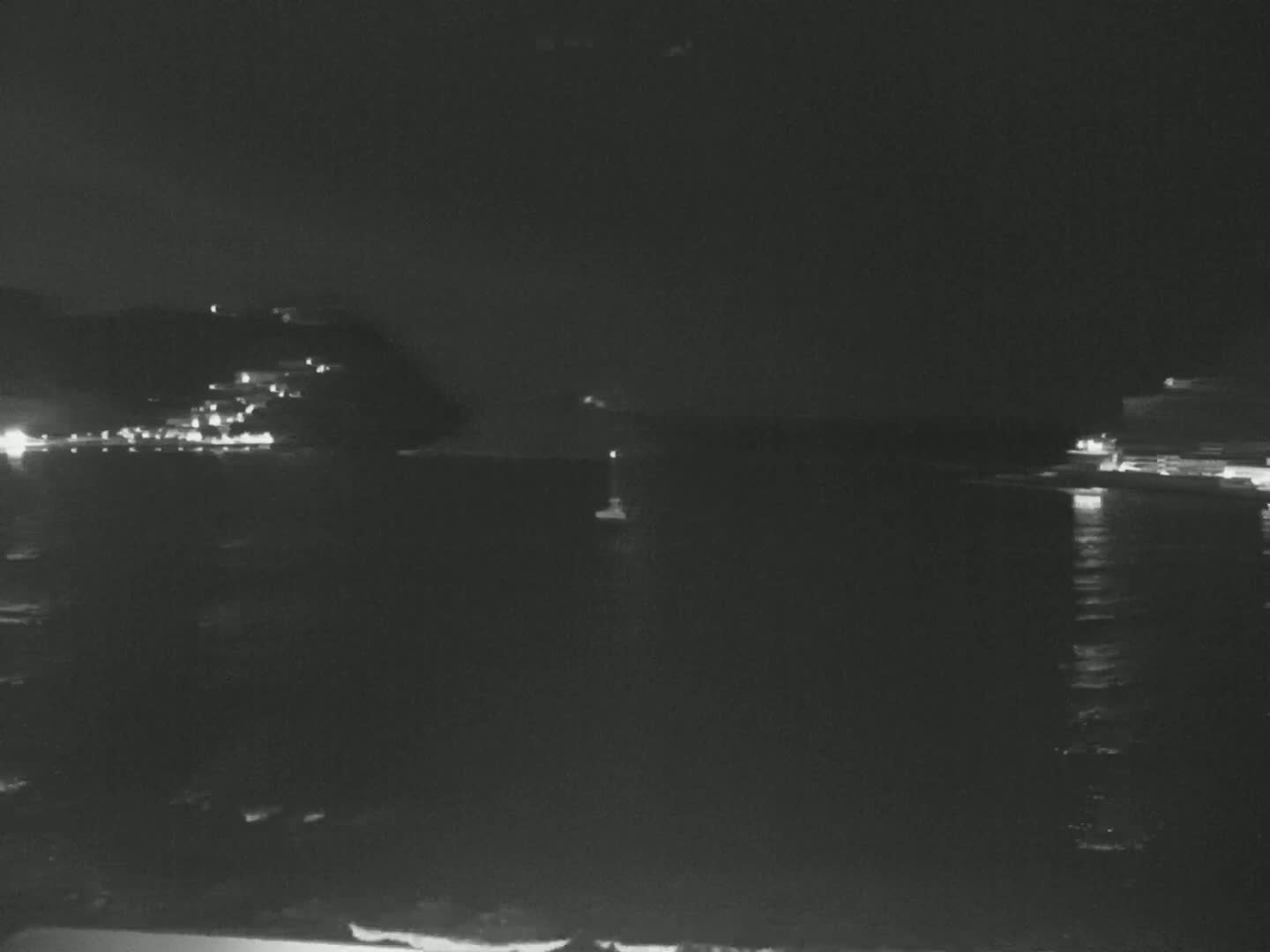 A nighttime, dark-grey seascape shows lights on a distant hillside and shoreline reflected in calm water.