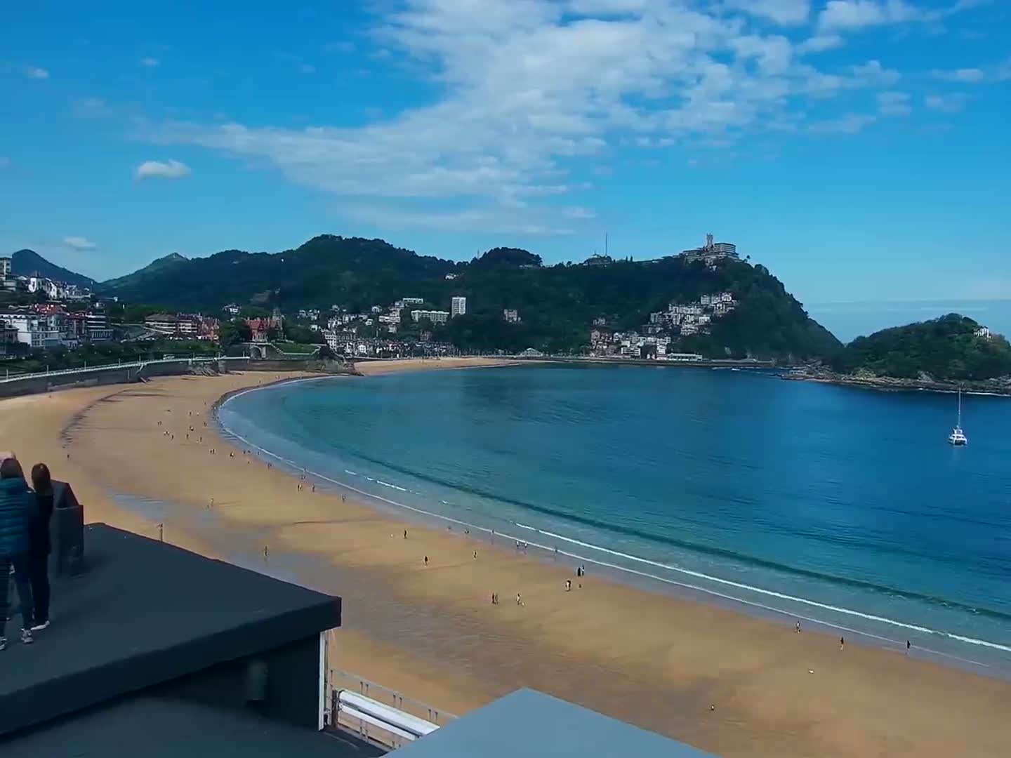 A mostly sunny day shows a curving sandy beach, calm blue water, and a town nestled against green hills, viewed from a rooftop.