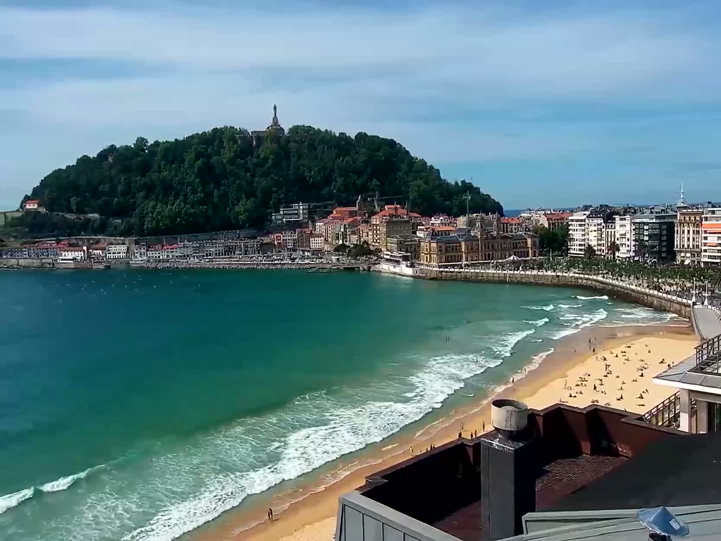 A sunny day reveals a coastal town nestled against a verdant hill, its sandy beach dotted with sunbathers and lapped by gentle waves.