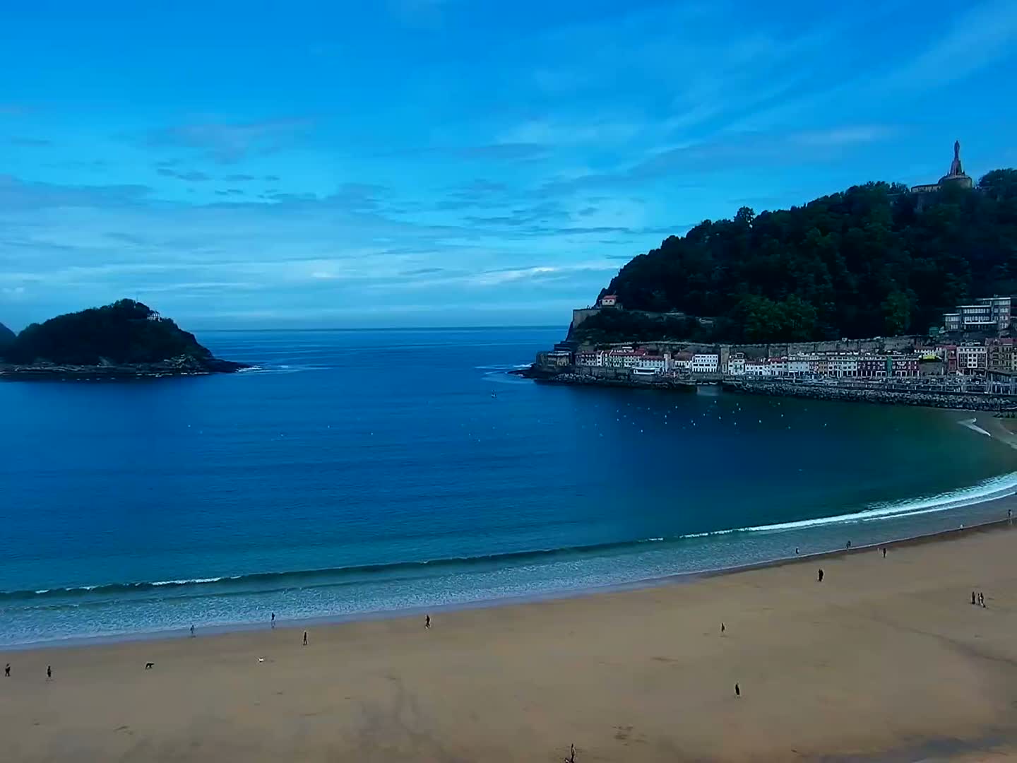 A sandy beach curves around a calm, blue bay, with a town nestled against a verdant hill in the background under a clear, blue sky.