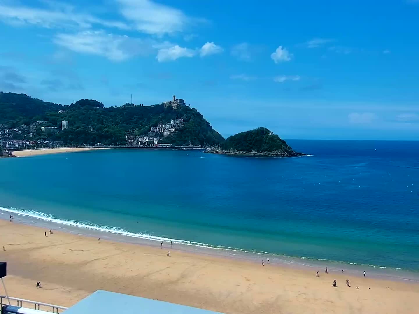 A sandy beach with a few people on it curves around a calm, clear blue ocean under a mostly sunny sky, with green hills and buildings visible in the distance.