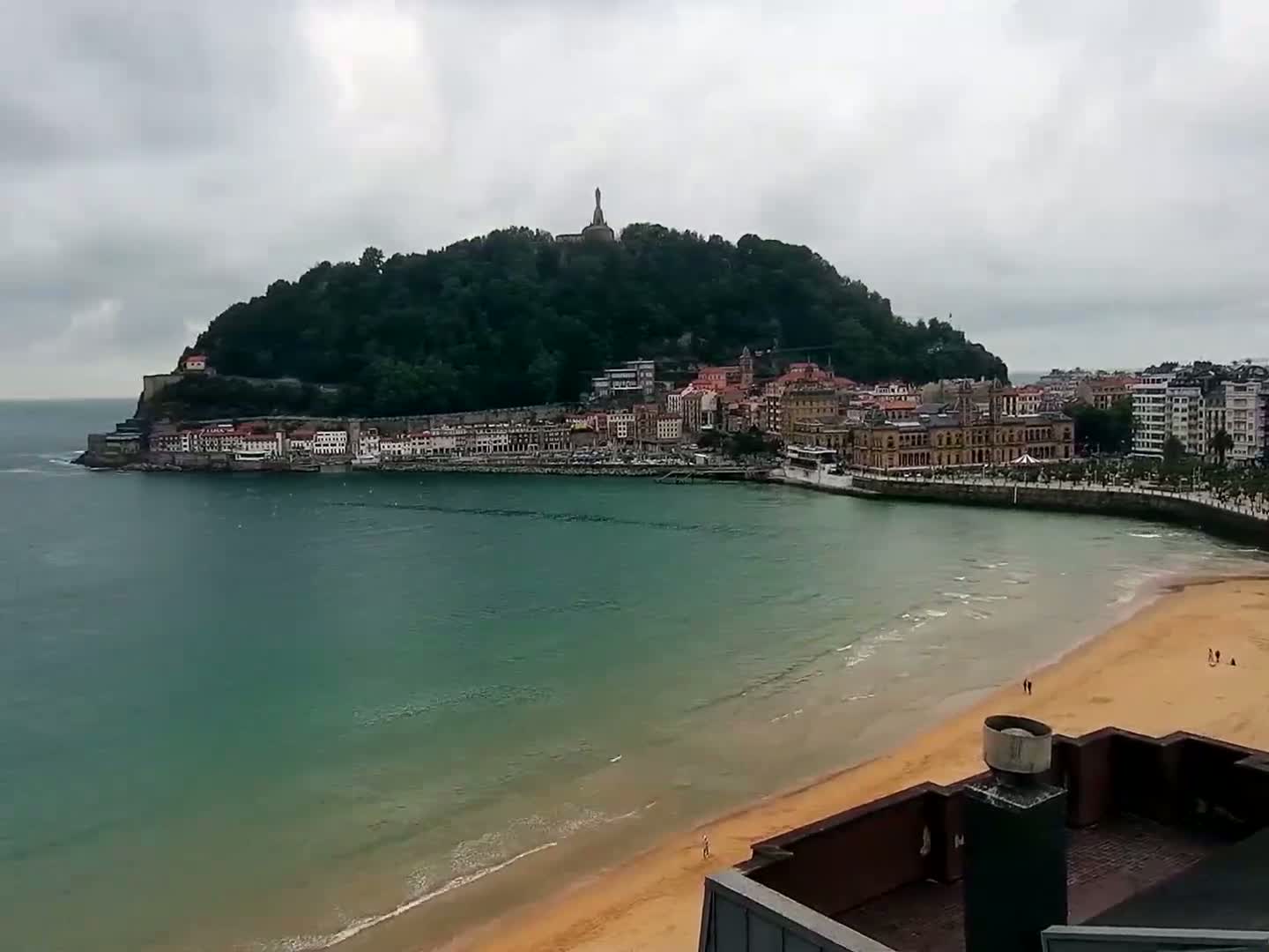 A coastal town nestled against a verdant hill, under a cloudy sky, features a sandy beach with a calm sea.