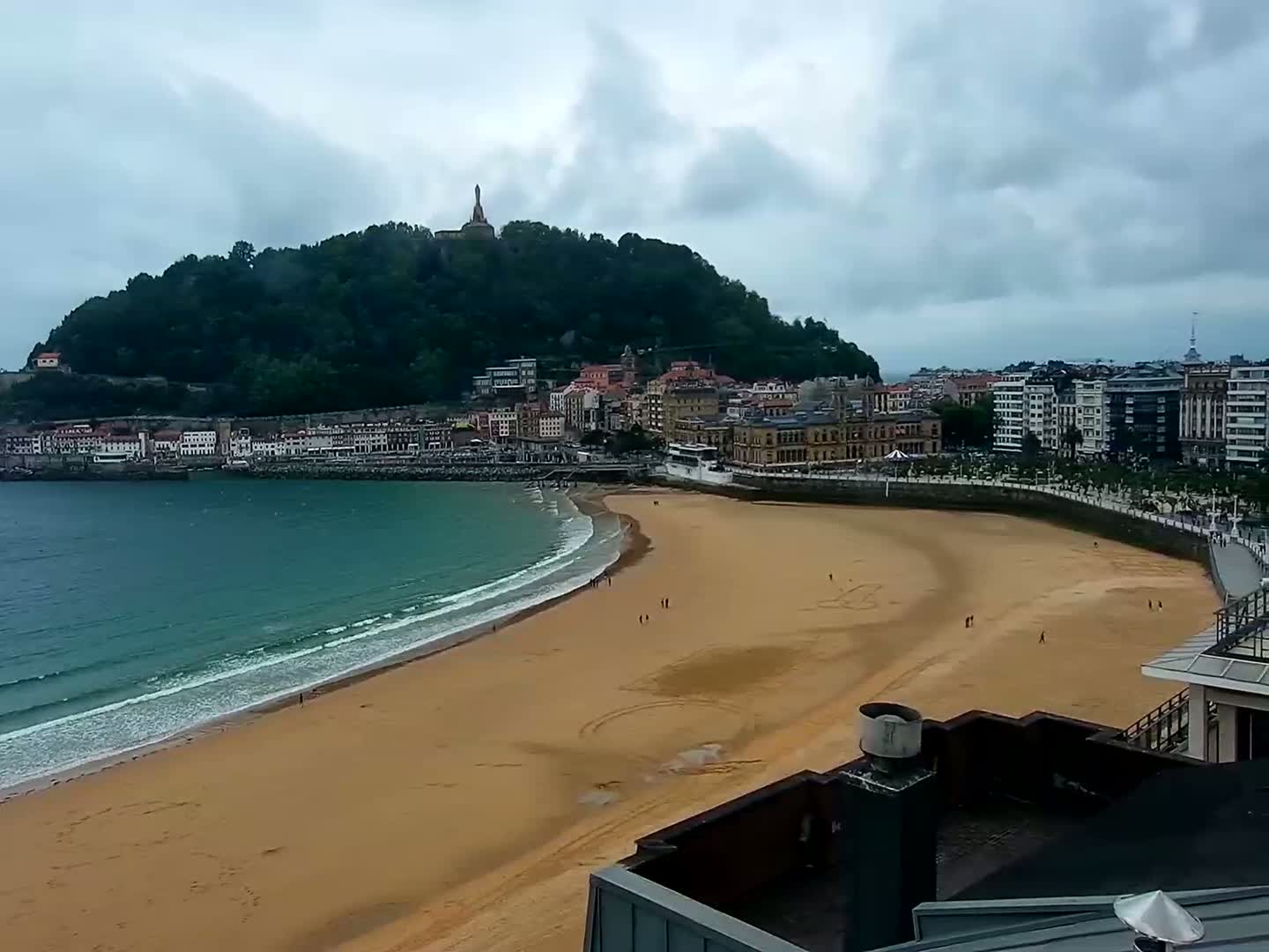 A sandy beach curves around a calm bay, next to a town nestled at the base of a green hill under an overcast sky.