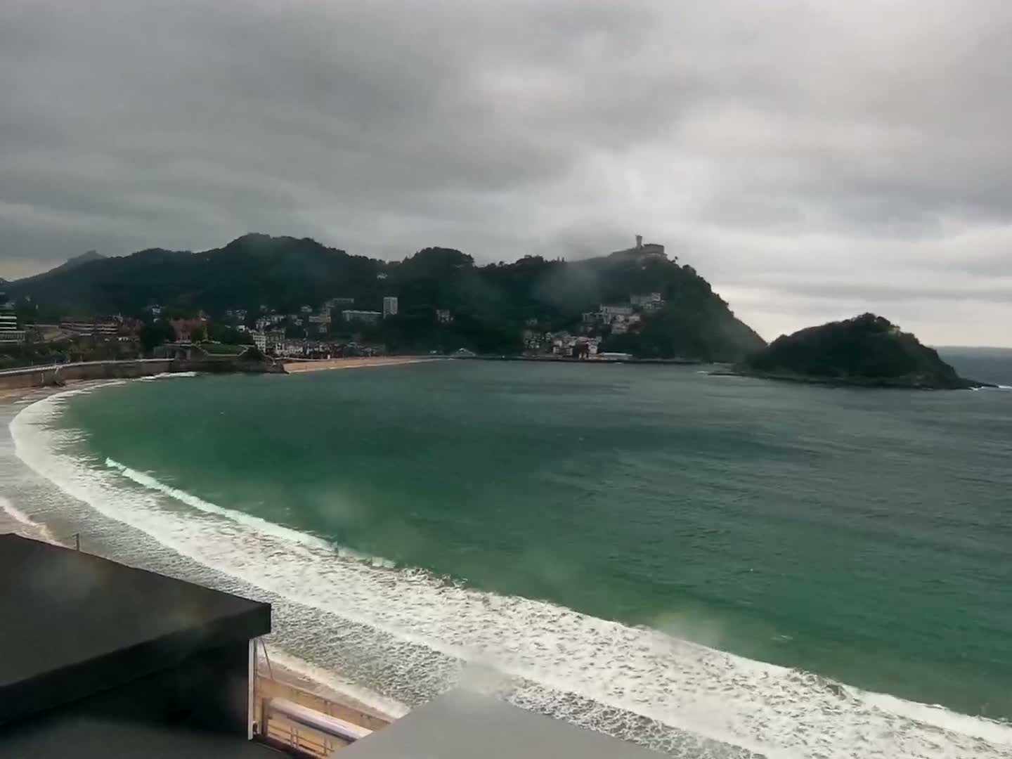A coastal town nestled beneath overcast skies sits on a curving bay with gentle waves lapping a sandy beach.