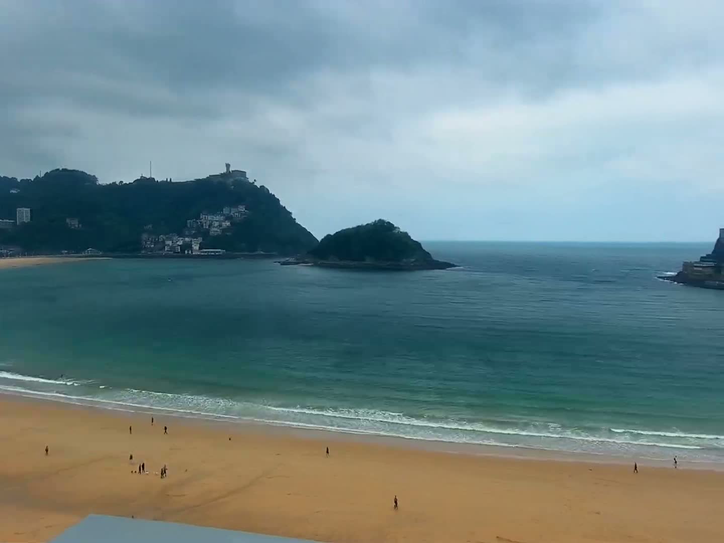 A sandy beach stretches below a hilltop town under an overcast sky, with a calm ocean separating it from a smaller island.