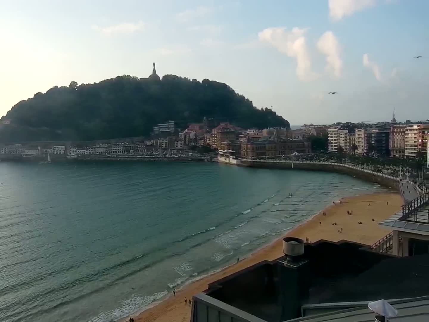 A sandy beach curves around a calm bay, with a town nestled at its edge beneath a verdant hill.