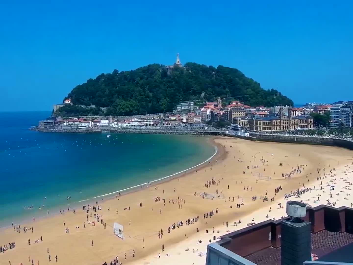 A sandy beach crowded with people curves around a bay, next to a town nestled at the base of a verdant, hilltop-crowned island under a clear blue sky.