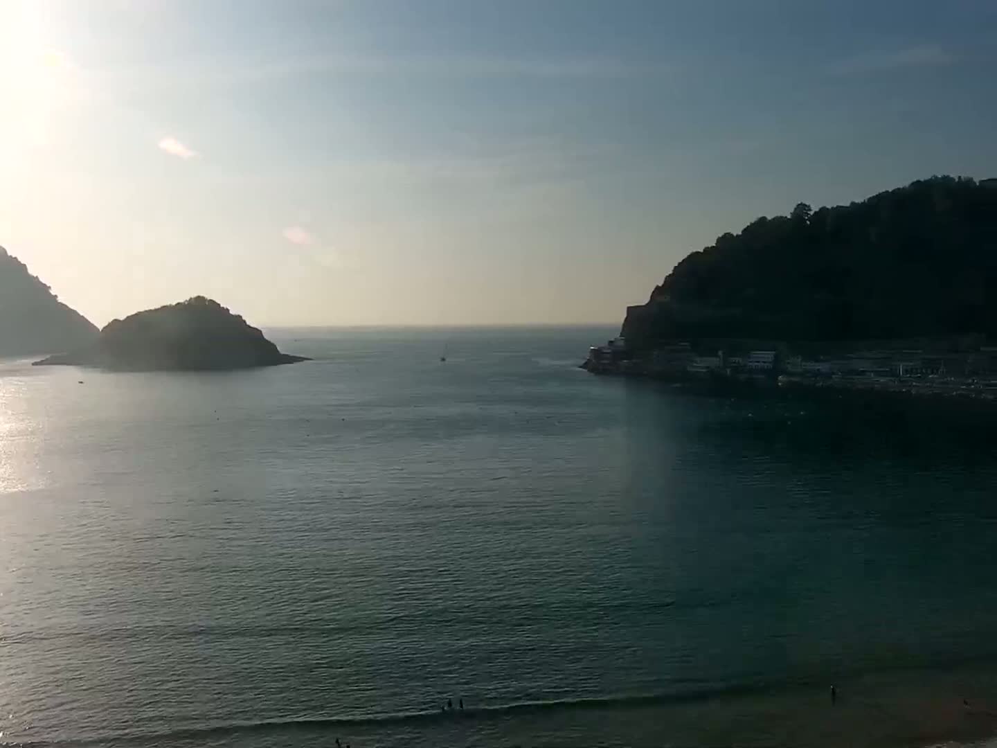 A calm, sunlit ocean bay is nestled between two dark, verdant hills, one of which has buildings along its shore.