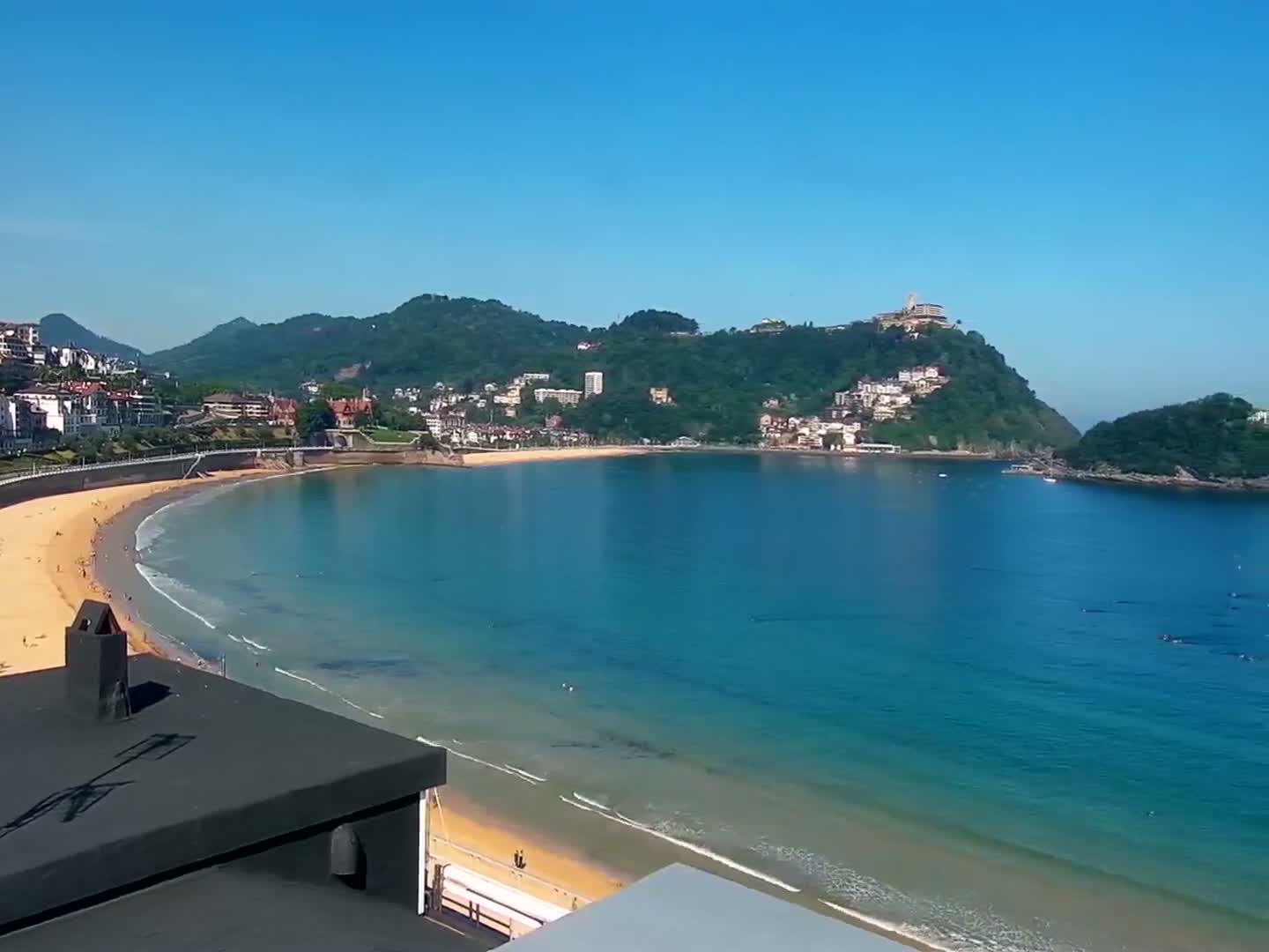 A curved sandy beach and calm, turquoise water are nestled in a bay surrounded by green hills and buildings under a clear blue sky.