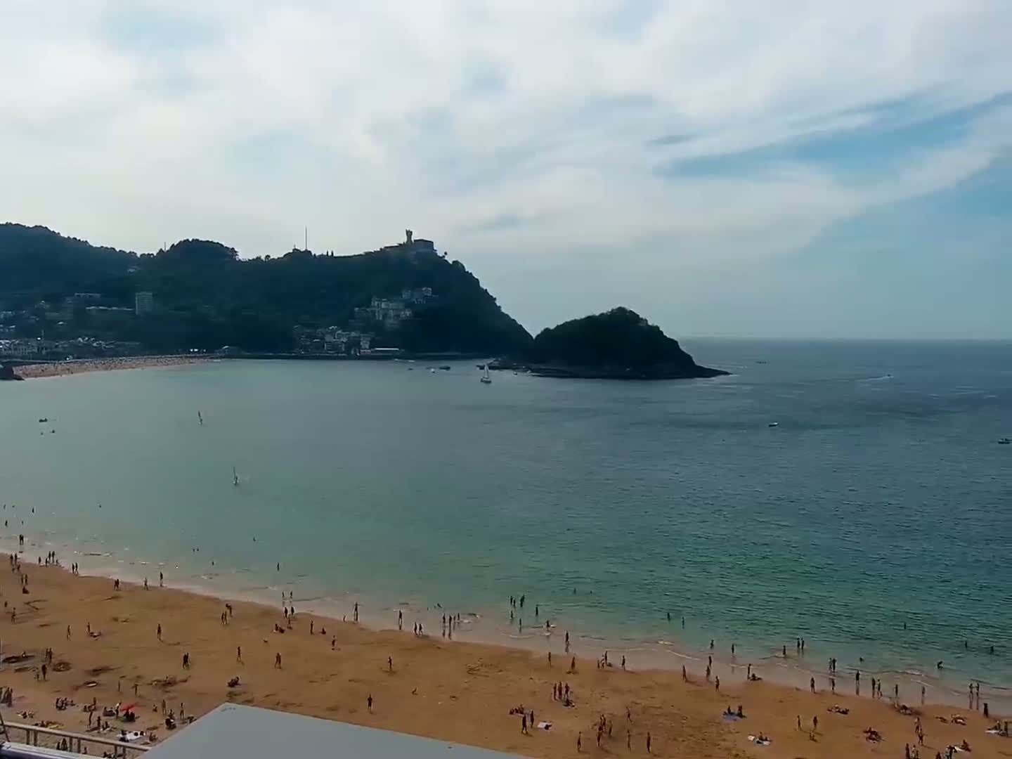 A sandy beach with many people on it is partially visible, with calm, teal water extending to a verdant hillside, featuring some buildings and a small island under a mostly cloudy sky.