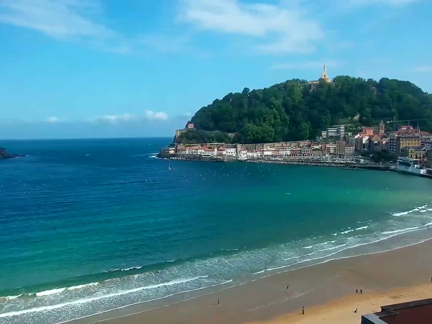 A sandy beach with gentle waves laps the shore of a turquoise bay, with a town nestled at the base of a green hill topped with a statue under a mostly sunny sky.