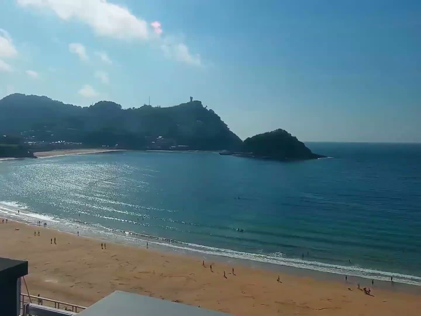 A sandy beach with a few people on it stretches along a calm, blue ocean beneath a mostly sunny sky and a line of hills.