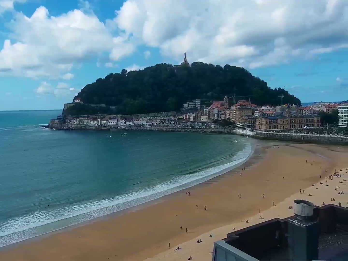 A sandy beach curves around a calm bay, with a town nestled at the base of a lush green hill under a partly cloudy sky.