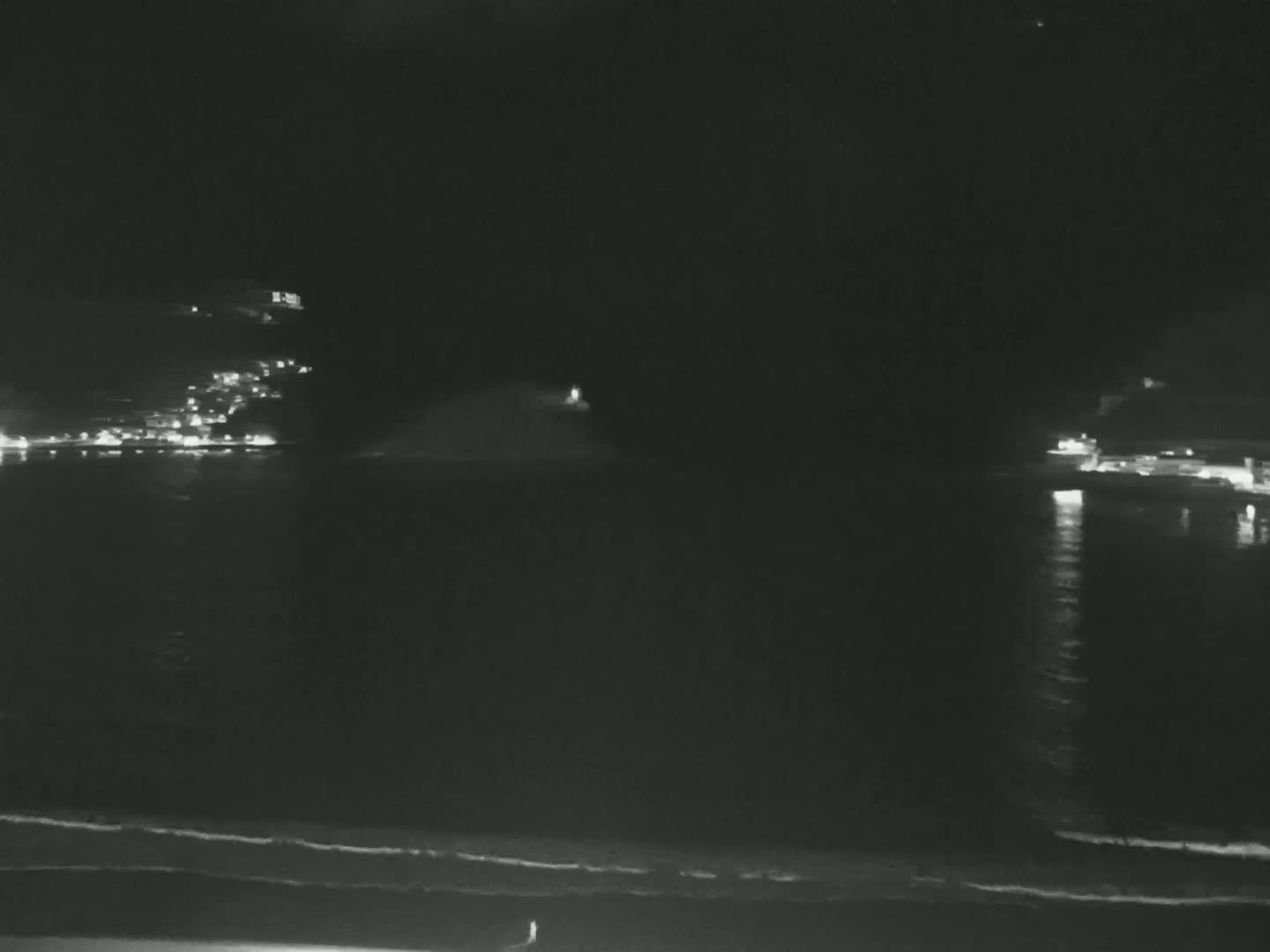 A dark, nighttime coastal scene shows a calm body of water reflecting the lights of buildings on the opposite shore, with a dark beach in the foreground.