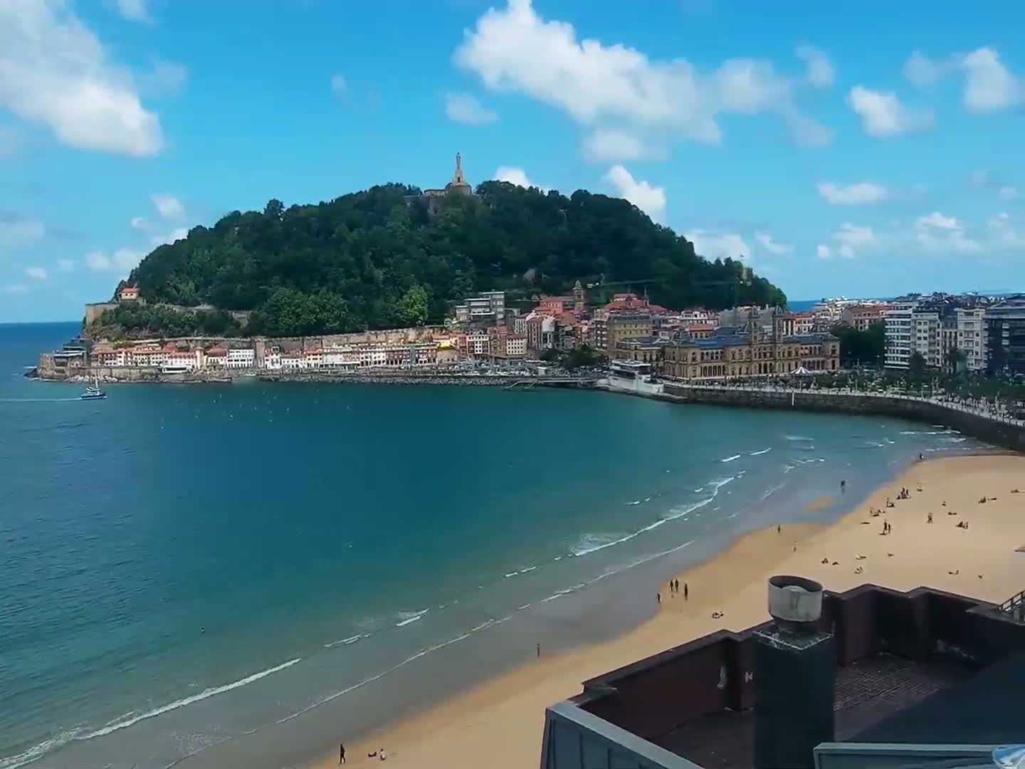 A sandy beach with gentle waves laps the shore beneath a blue sky dotted with clouds, in front of a town nestled at the base of a green hill topped with a monument.