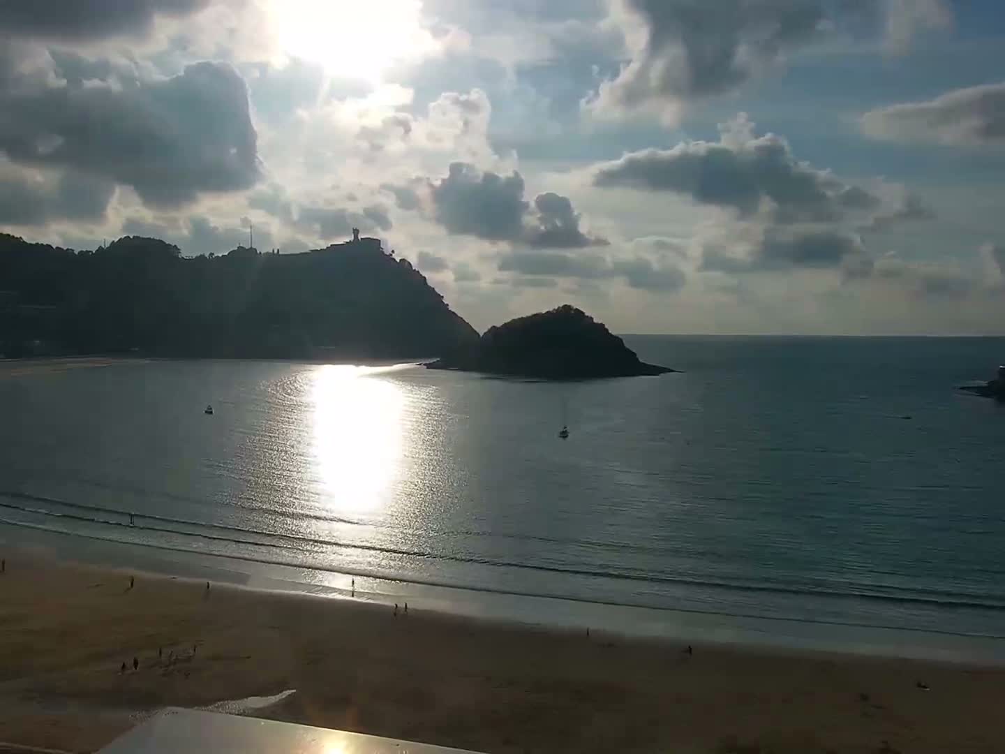 A sandy beach with a few people on it stretches out to calm water reflecting sunlight under a partly cloudy sky, with hills and a small island in the background.