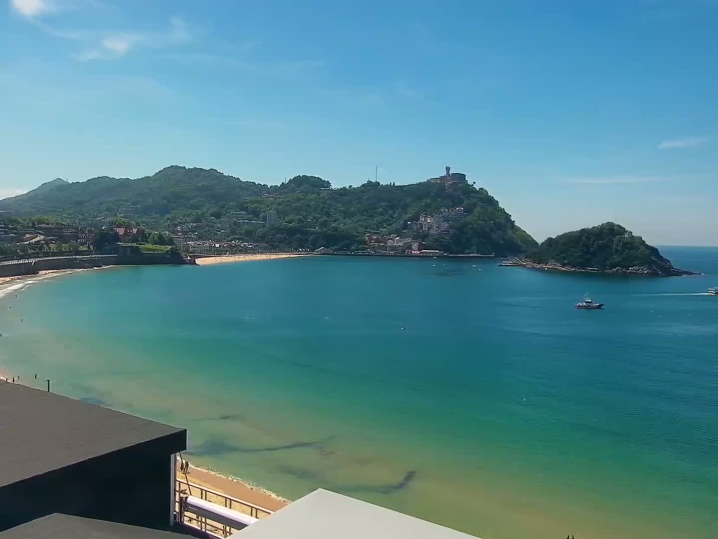 A sandy beach curves around a bay of clear, teal water under a bright, sunny sky, with a green hill topped by buildings in the background.