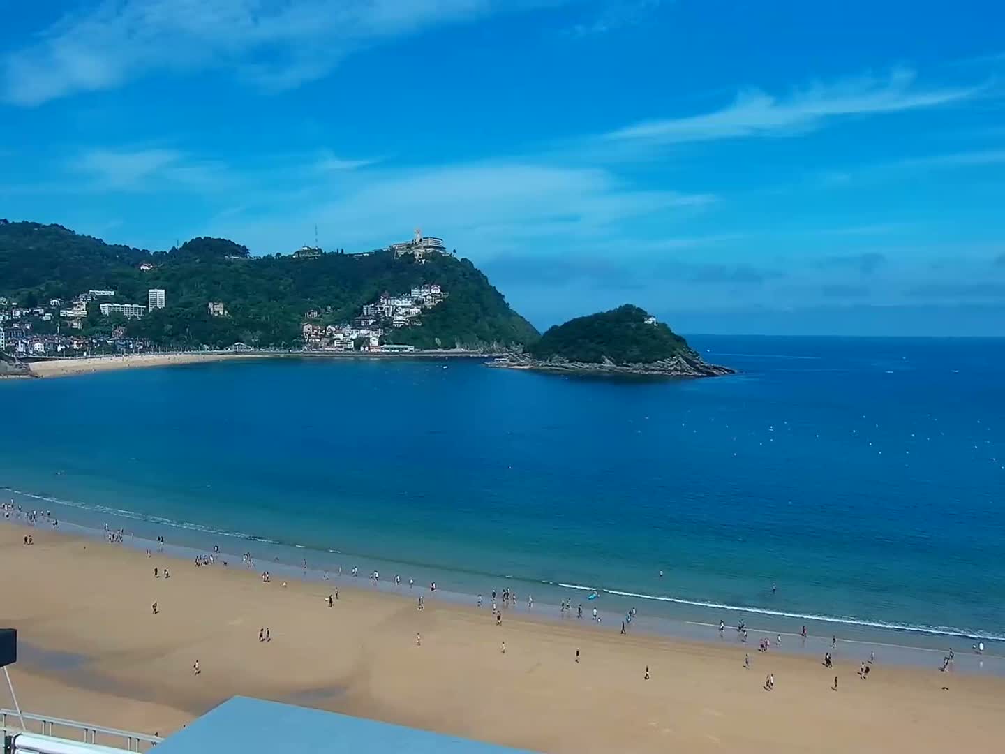 A sandy beach with scattered people is situated on a bay next to a lush green hill under a mostly clear blue sky.