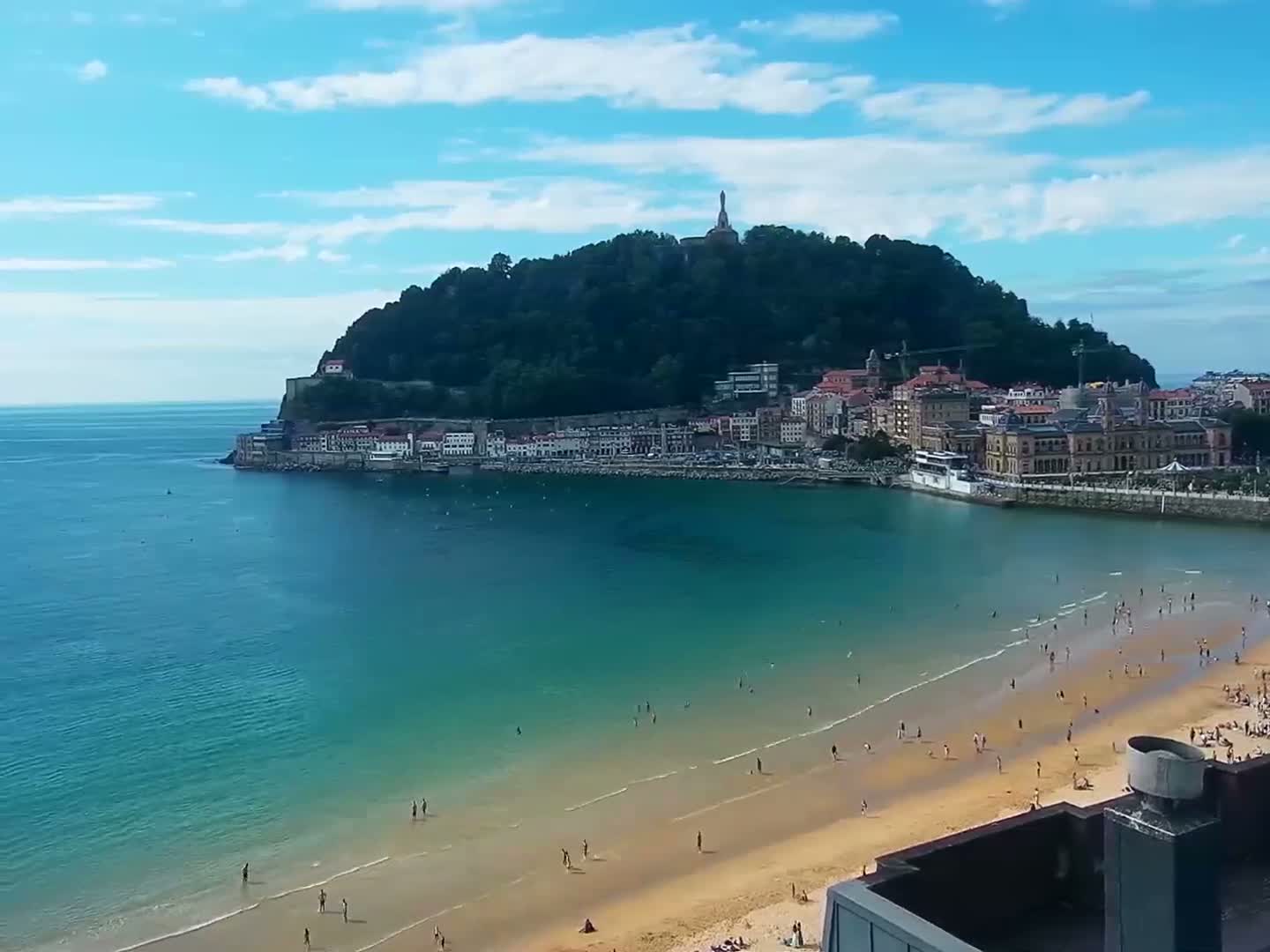 A sandy beach with many people on it curves around a bay toward a town situated at the base of a green hill under a mostly clear blue sky.