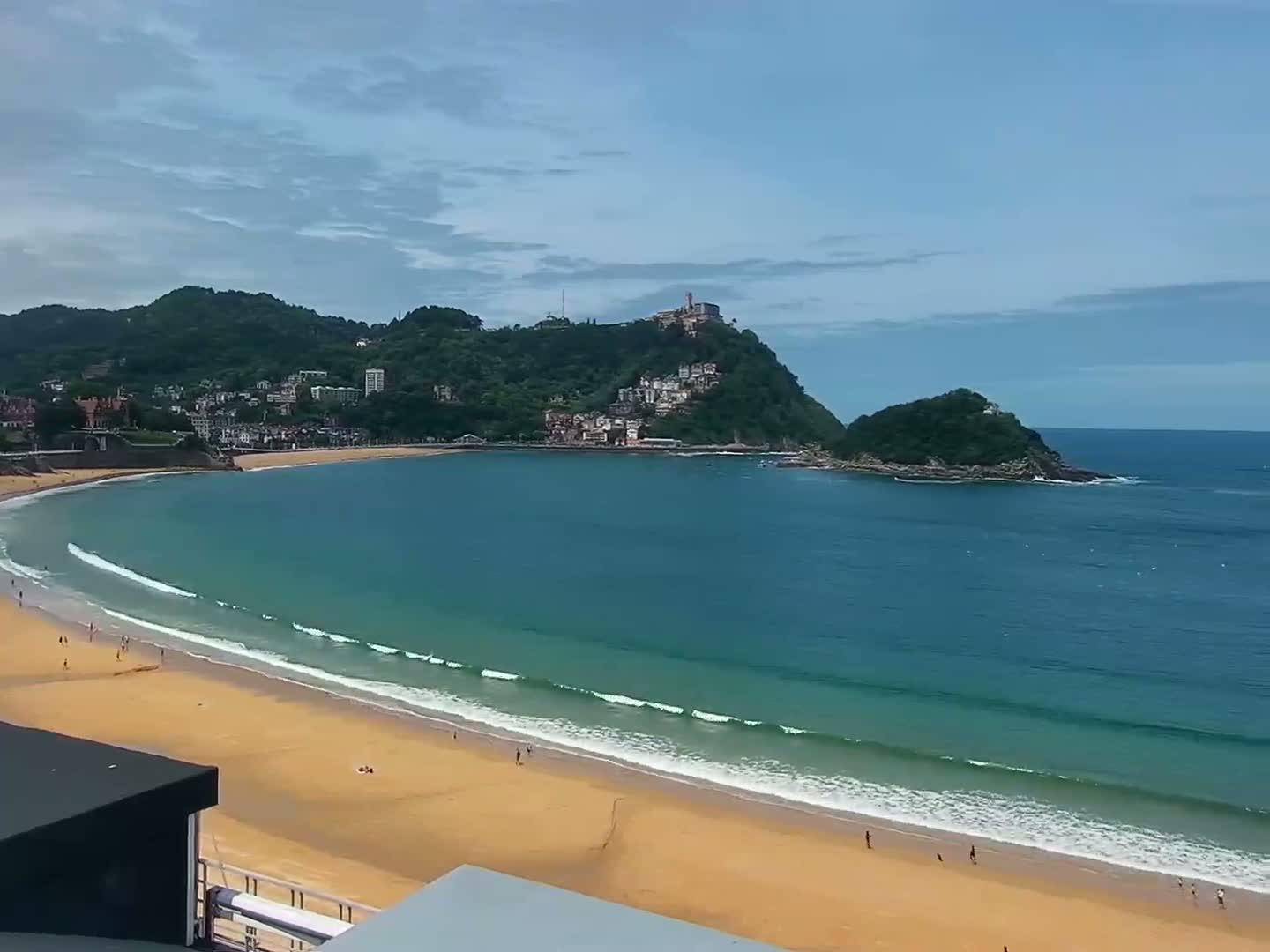 A sandy beach curves around a bay of calm, turquoise water, backed by green hills and a town nestled against a hill under a mostly clear sky.