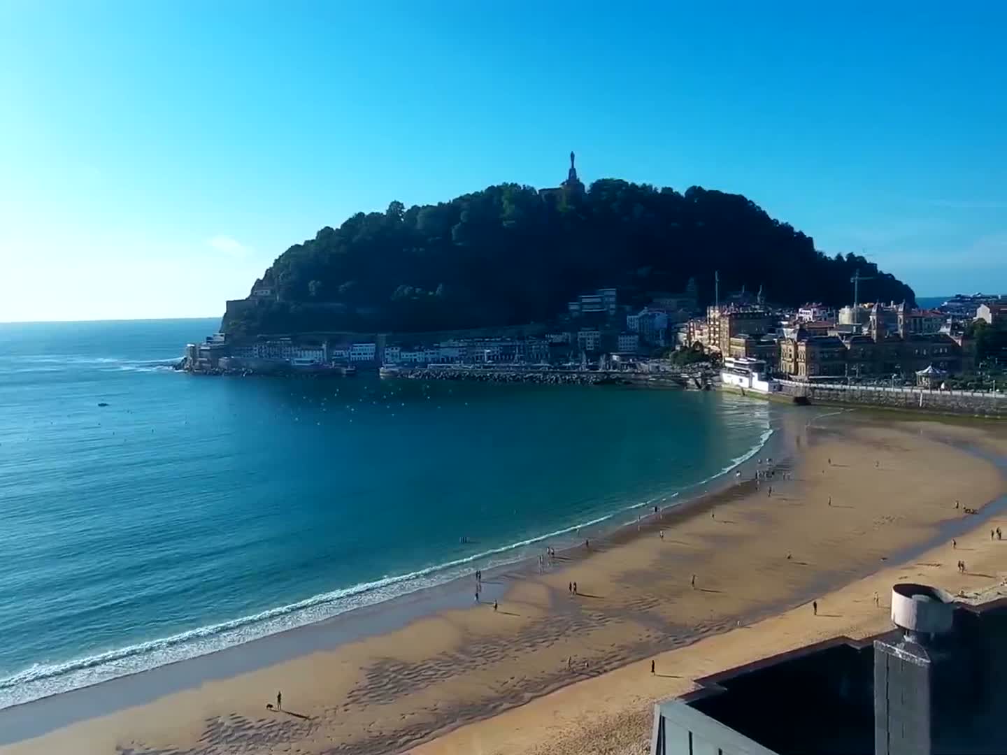 A sandy beach curves around a calm bay, with a town nestled at the base of a verdant hill under a clear blue sky.