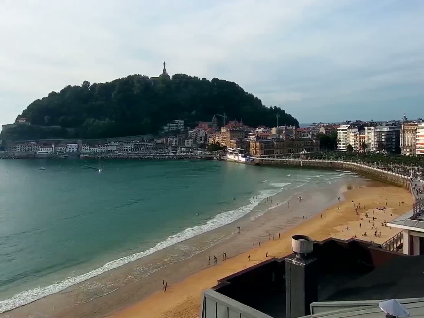 A sandy beach with people sunbathing and swimming is seen from an elevated vantage point, with a calm ocean and a town nestled against a green hill in the background under a mostly cloudy sky.