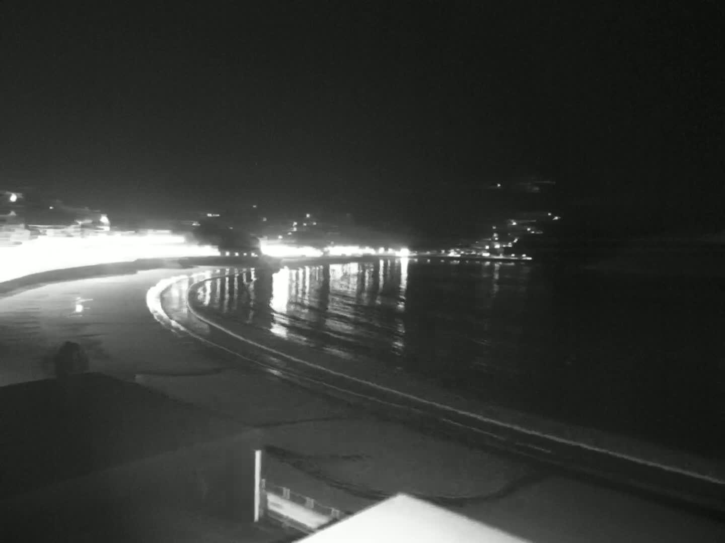 A nighttime, wet beach curves along a calm body of water reflecting distant lights.