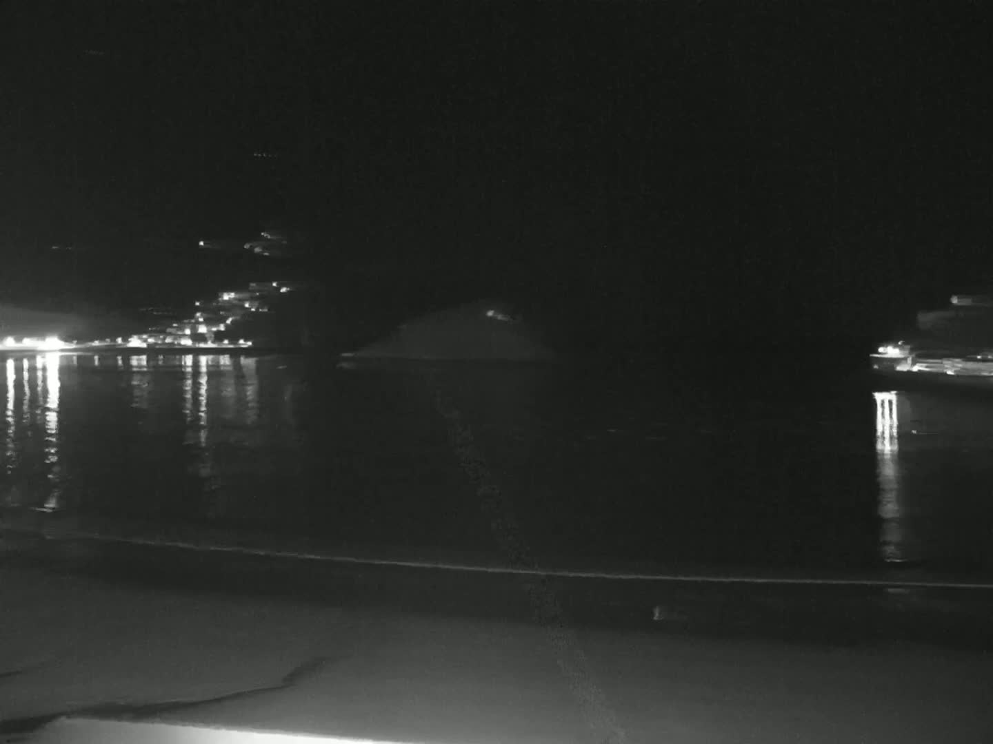 A nighttime view of a dark body of water reflecting lights from a distant town, with a sandy beach in the foreground under a clear sky.