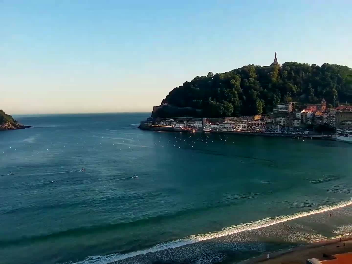 A coastal town nestled beneath a verdant hill, topped with a monument, overlooks a calm, blue ocean with gentle waves lapping a sandy beach under a clear, bright sky.