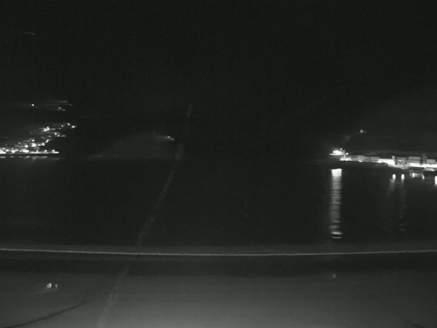 A nighttime long shot shows a dark body of water reflecting lights from buildings on the far shore, with a dark road in the foreground.