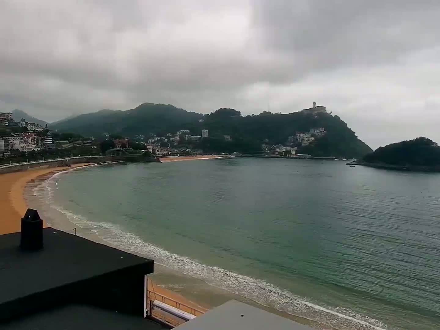 A sandy beach curves into a calm bay under a cloudy sky, with green hills and buildings lining the shore.