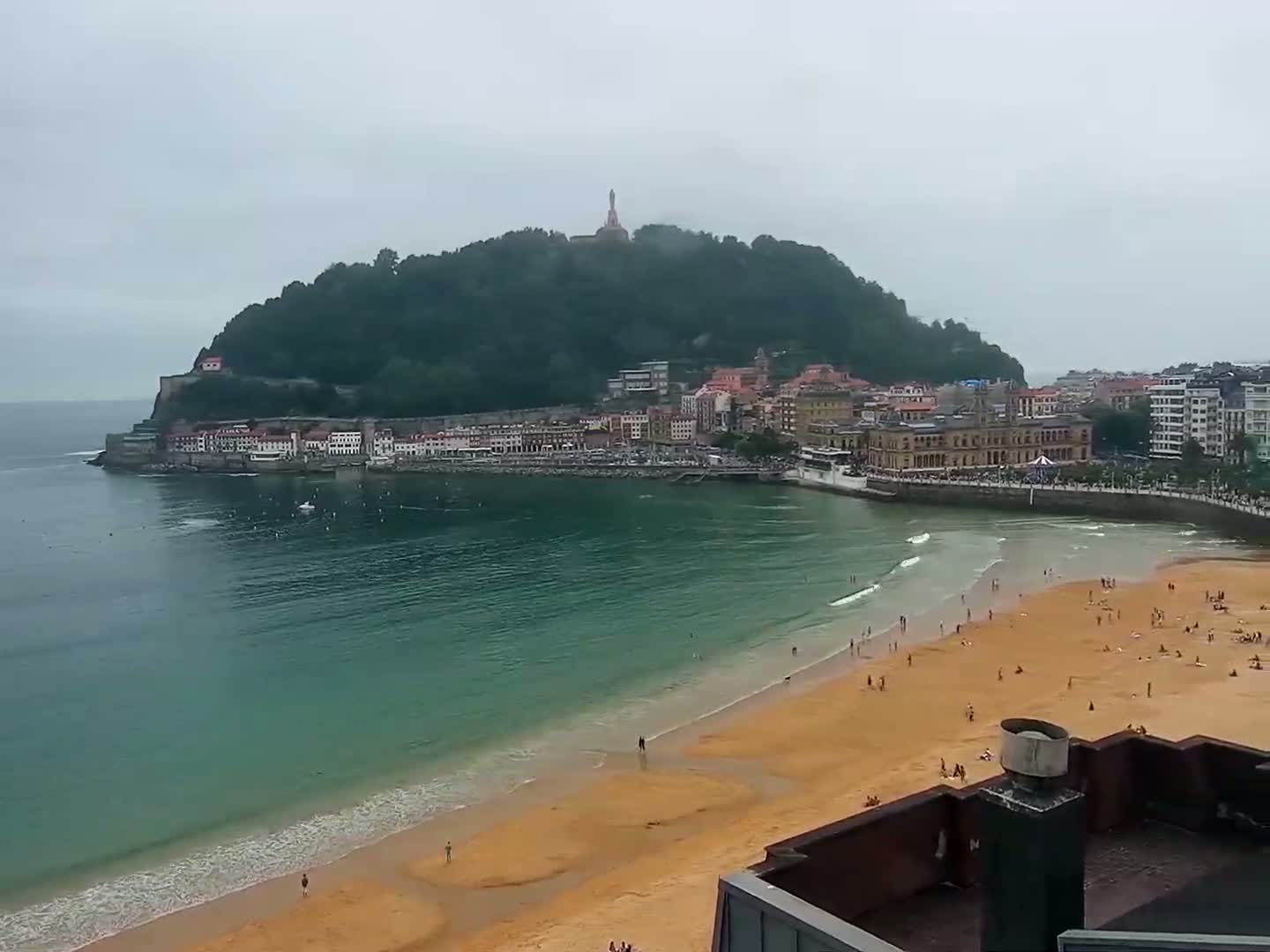 A sandy beach with people on it is situated next to a town nestled at the base of a verdant, hilltop with a monument, all under a cloudy sky.