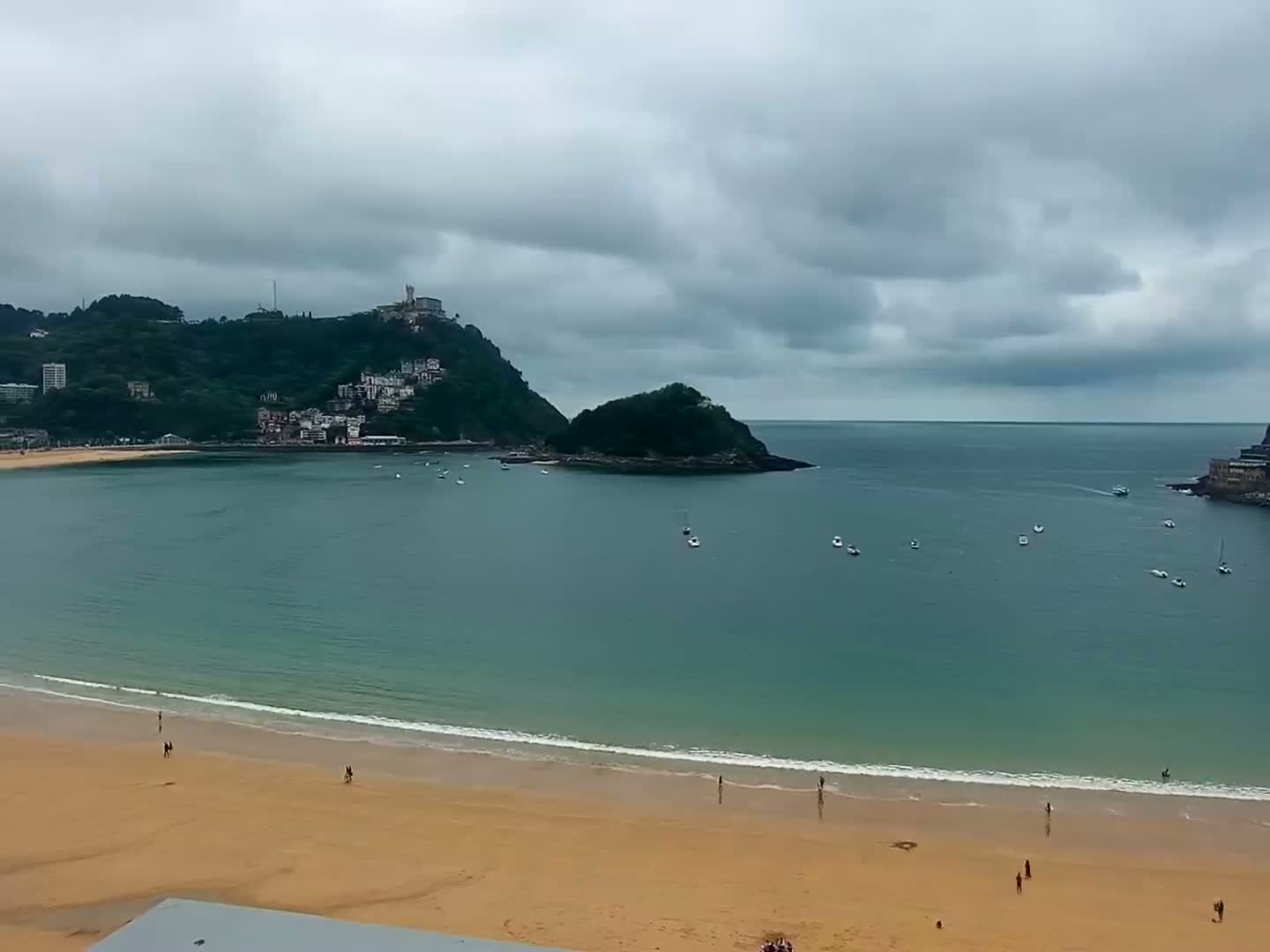 A sandy beach with a few people on it stretches along a calm bay under a cloudy sky, with a green hill topped with buildings in the background.